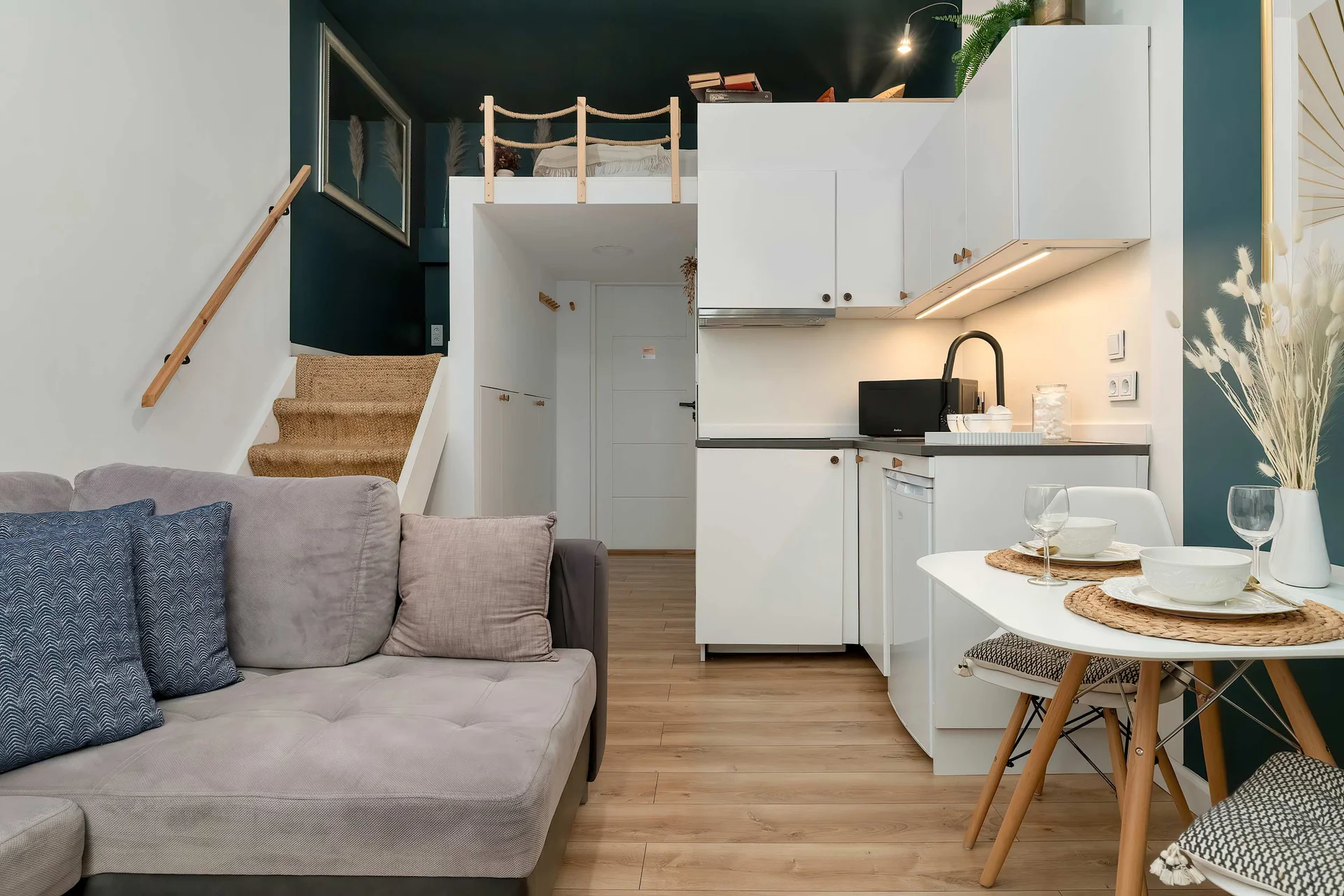 A bright open-plan interior combining a small living area, dining space, and kitchenette with warm wooden flooring throughout.