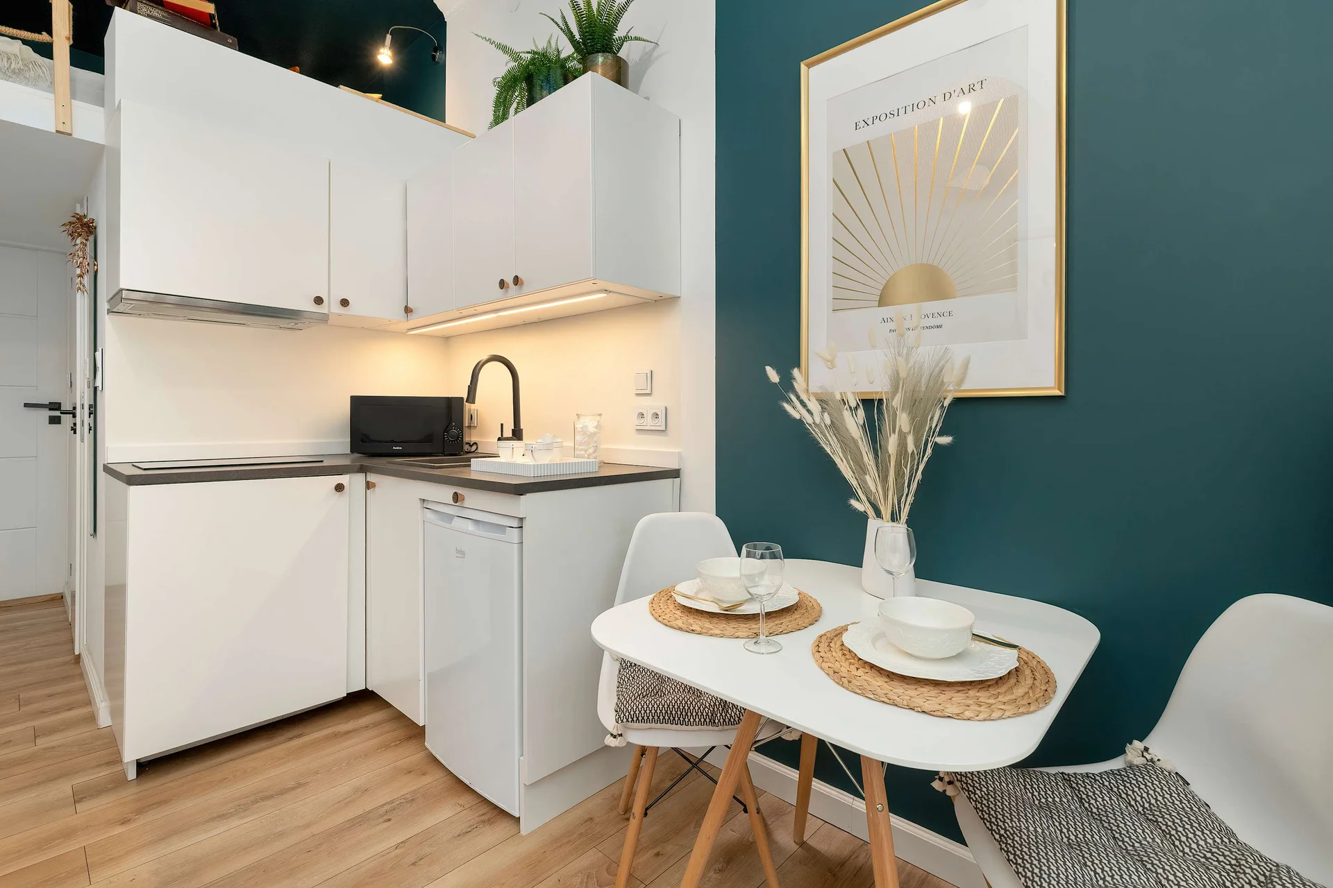 A compact dining area next to a white kitchenette with sleek cabinetry. Two place settings are arranged on a round table against a dark teal accent wall.