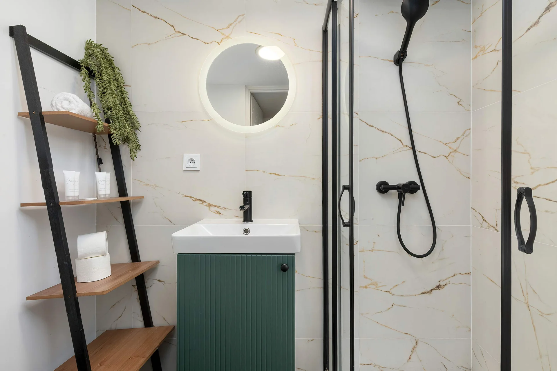 A stylish bathroom with white marble-patterned walls and black fixtures. A round illuminated mirror hangs above a green vanity cabinet, while a ladder shelf provides storage.