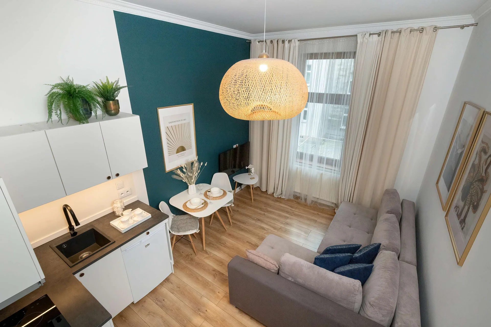 A view of the living area from another angle, showing a gray sofa, dining area, and part of the bathroom entrance. Pendant lighting and warm wood tones add a welcoming feel.