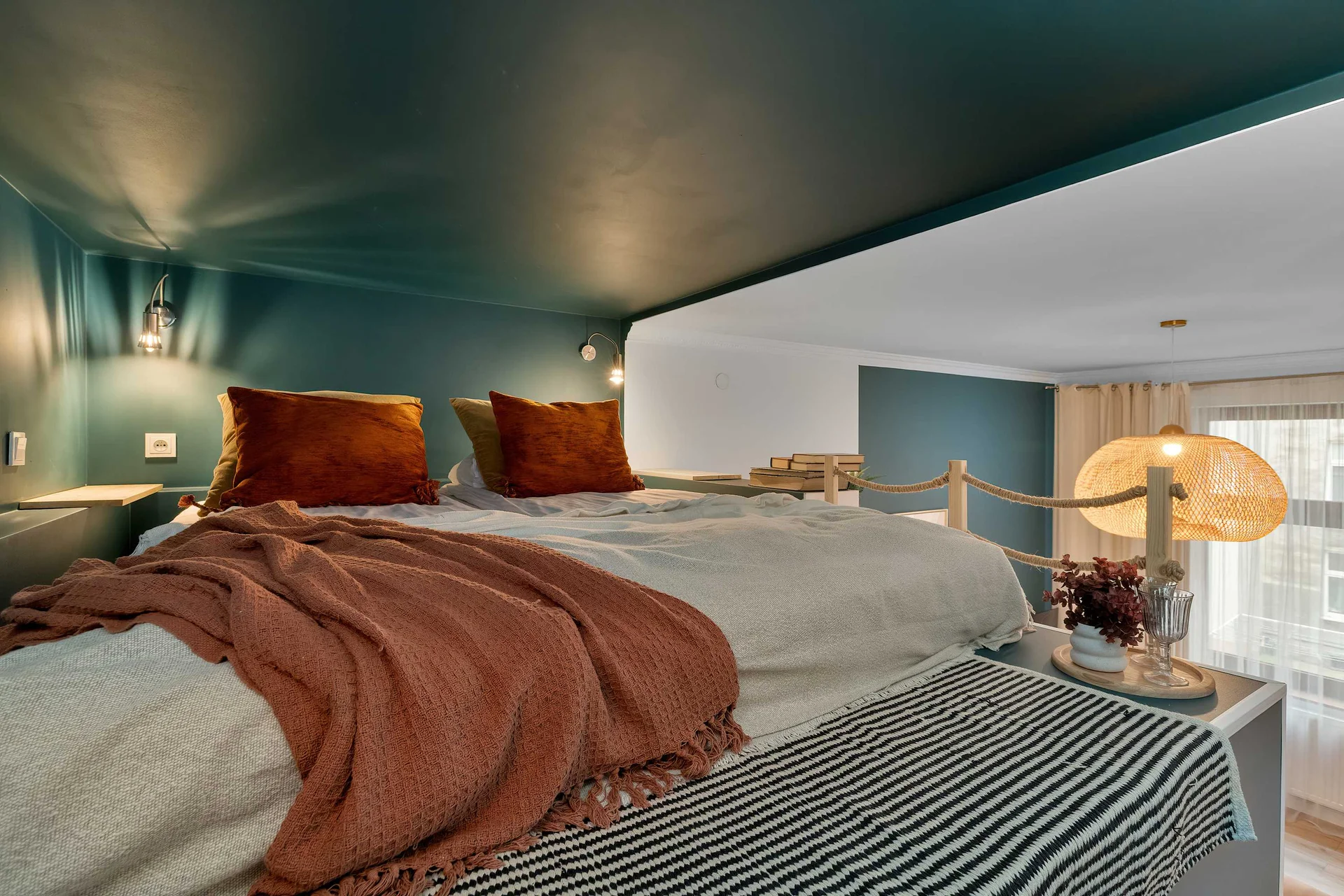 A cozy sleeping area featuring a large bed dressed with white linens and a rust-colored throw blanket. Soft lighting and deep teal walls create a modern, intimate atmosphere.