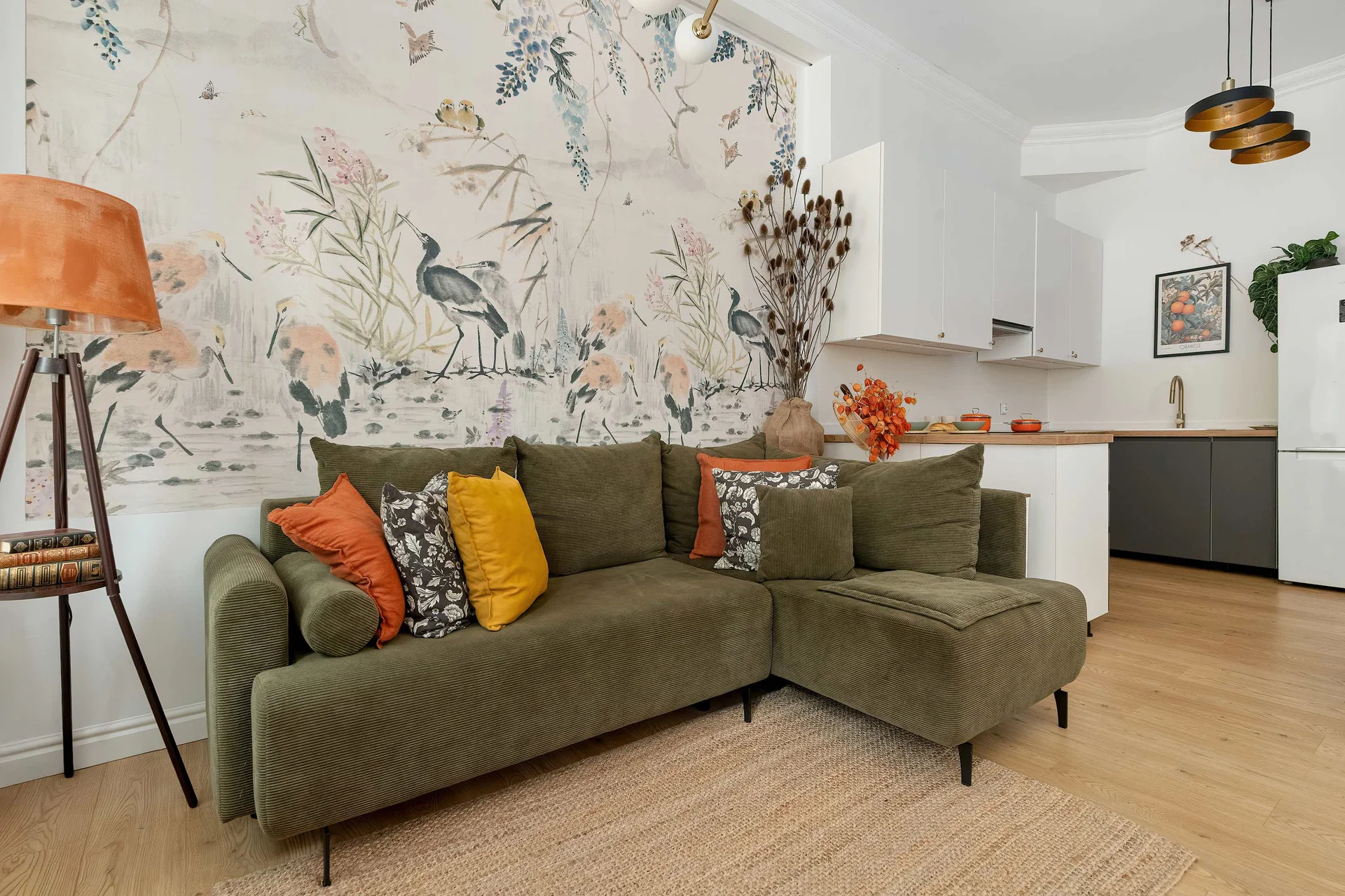 A welcoming lounge with an olive-green sofa, colorful cushions, botanical wallpaper, and a floor lamp beside the seating area.