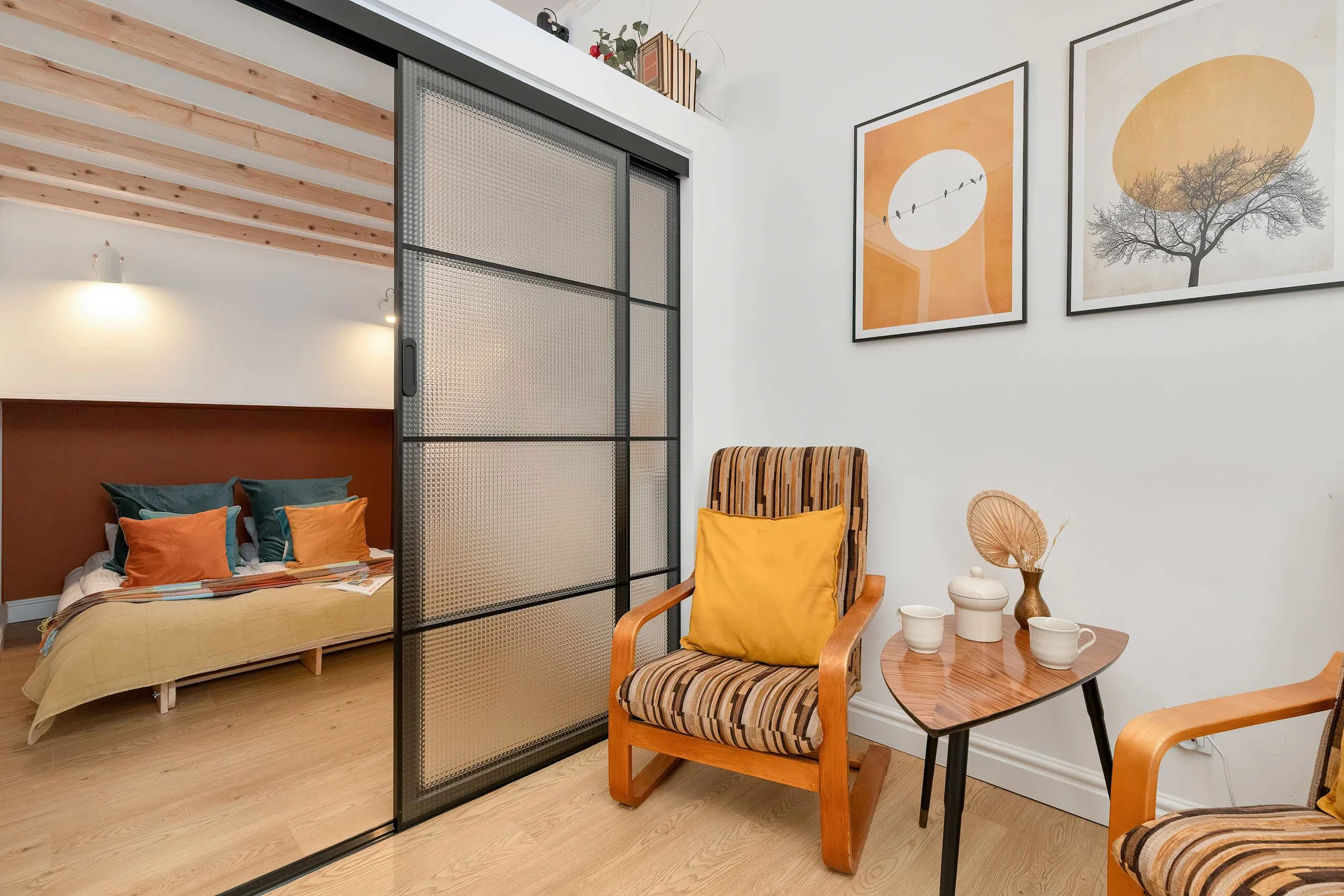 A stylish bedroom area, a mustard-yellow armchair, and framed artwork displayed above a small side table.