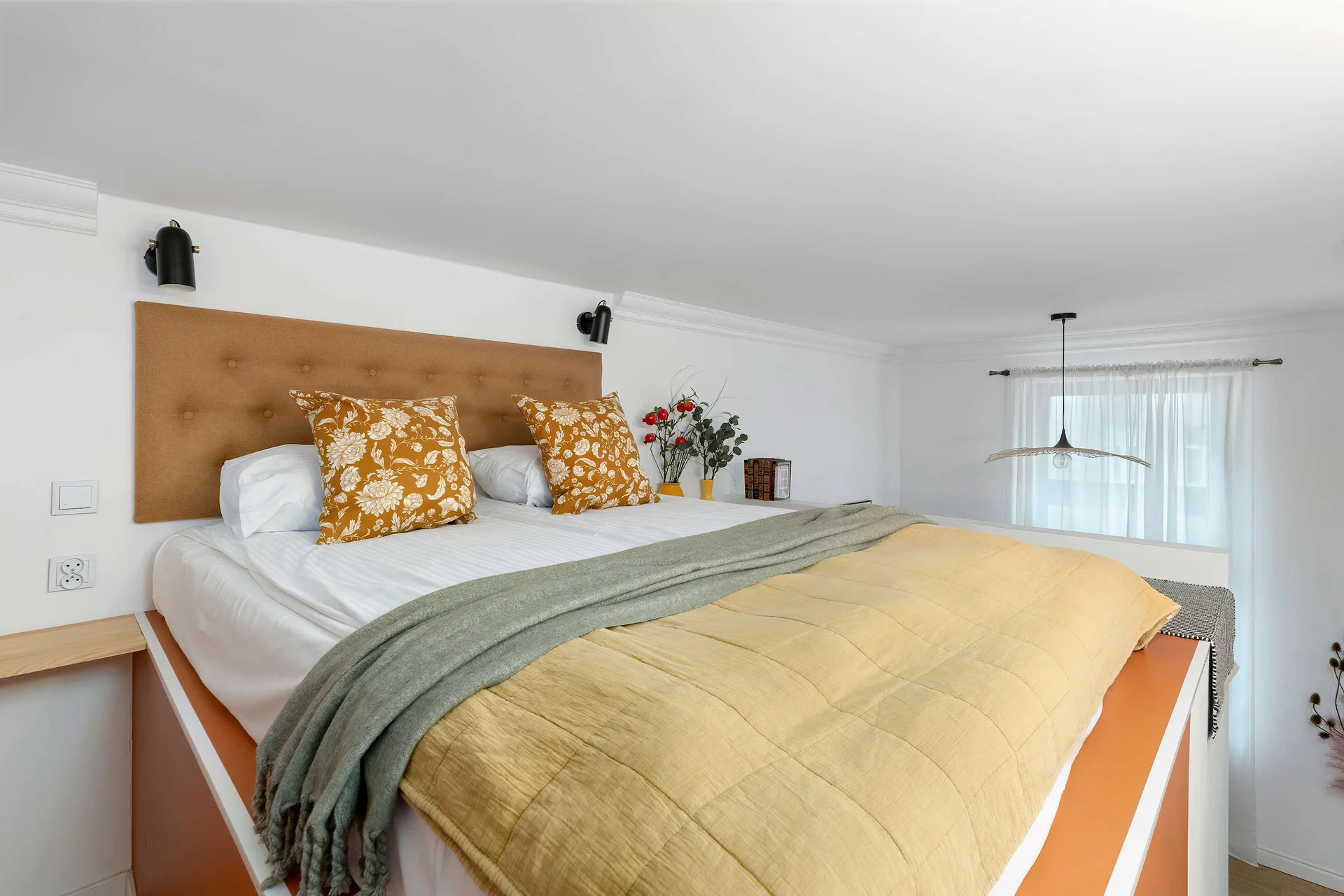 A comfortable bedroom area featuring a double bed with soft green bedding, mustard accents, and a wooden headboard decorated with floral details.