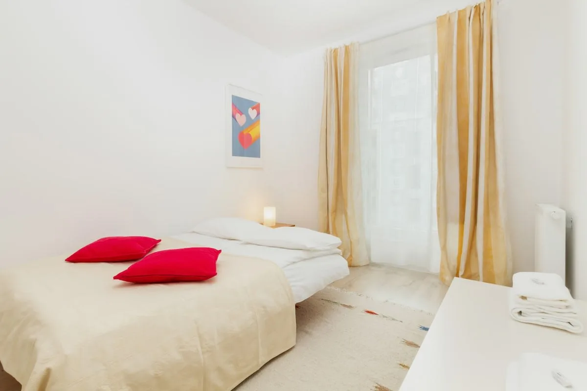 A cozy bedroom with a double bed, white bedding, and red accent pillows. Natural light fills the room, complemented by a bedside table and lamp.