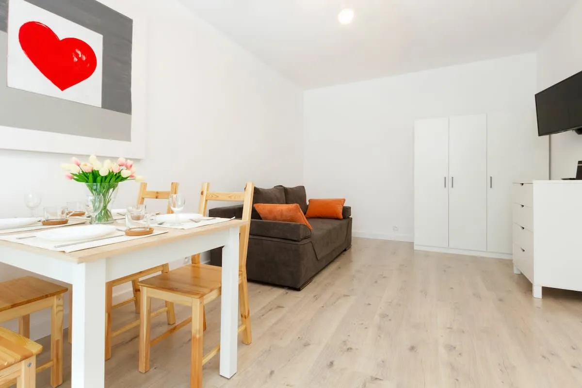 A bright, modern living room featuring a dining table with wooden chairs, and a comfortable sofa in the background. The decor is minimalistic with light wooden floors and white walls.