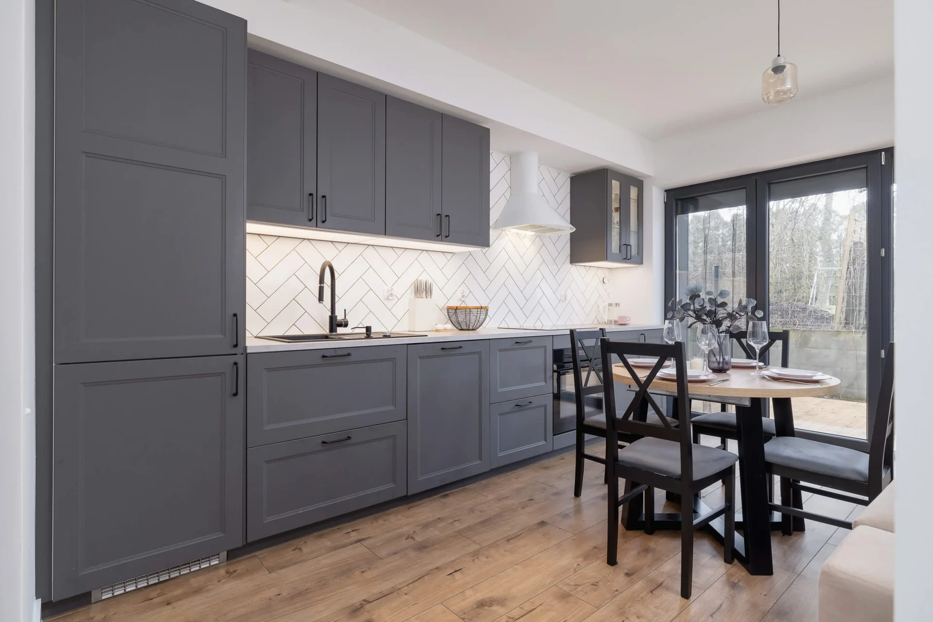 A contemporary kitchen with dark gray cabinets, a sleek black dining table, and large windows offering a view of the outdoor patio.