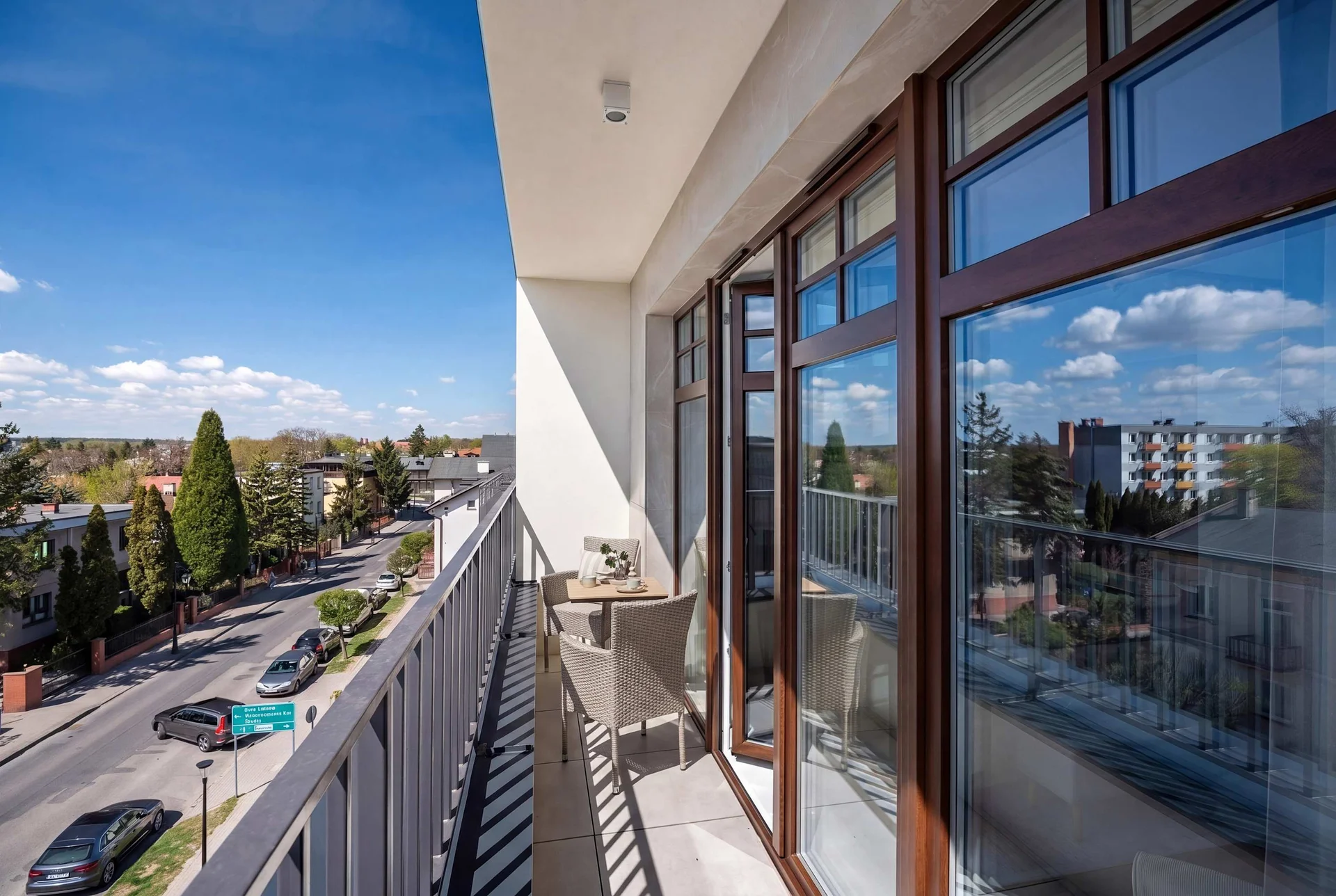 Looking along the balcony, the view captures the urban landscape and the building's modern architecture. The wooden-framed glass doors reflect the blue sky and white clouds.