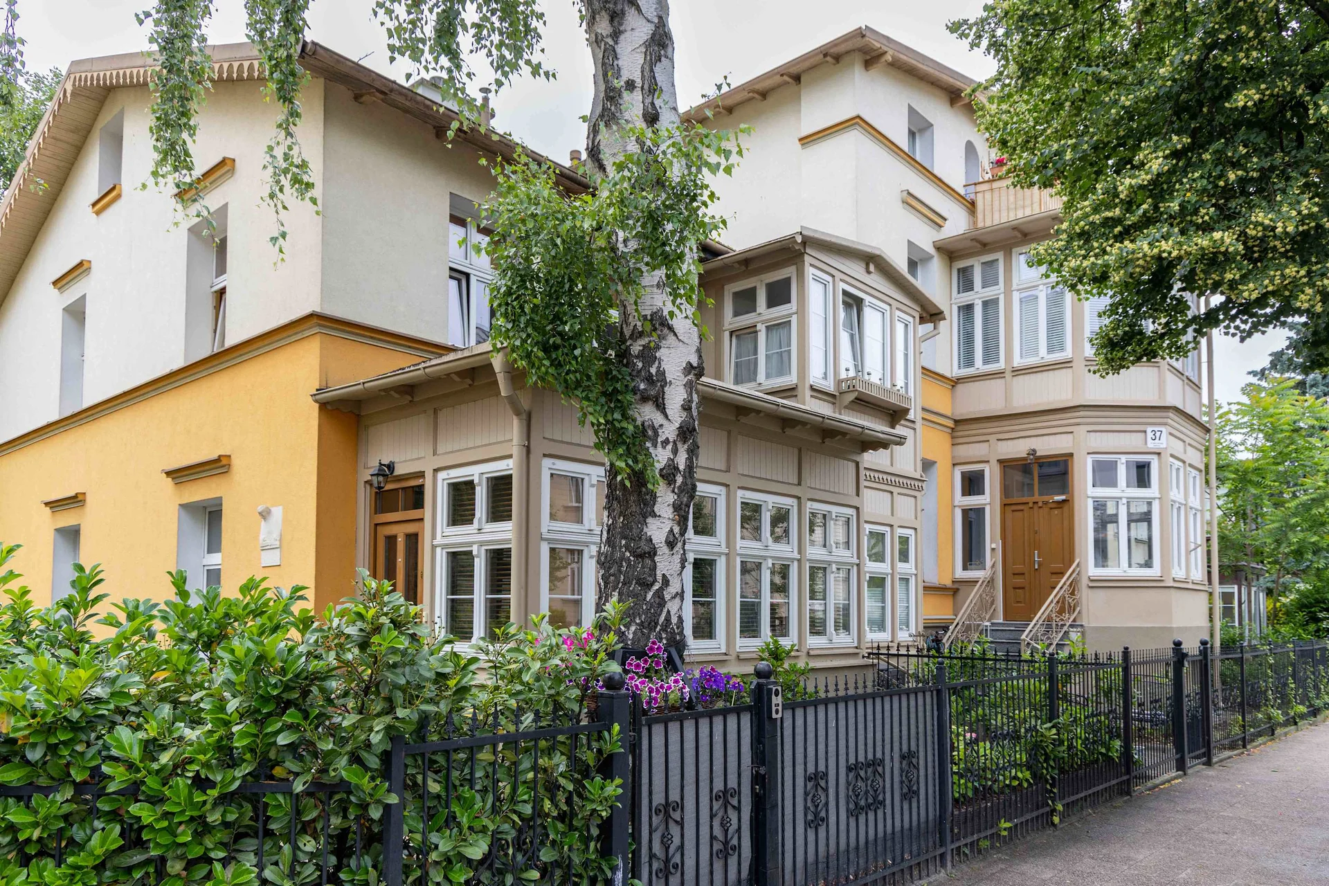 A charming residential building surrounded by trees and greenery. The entrance is fenced and well maintained.