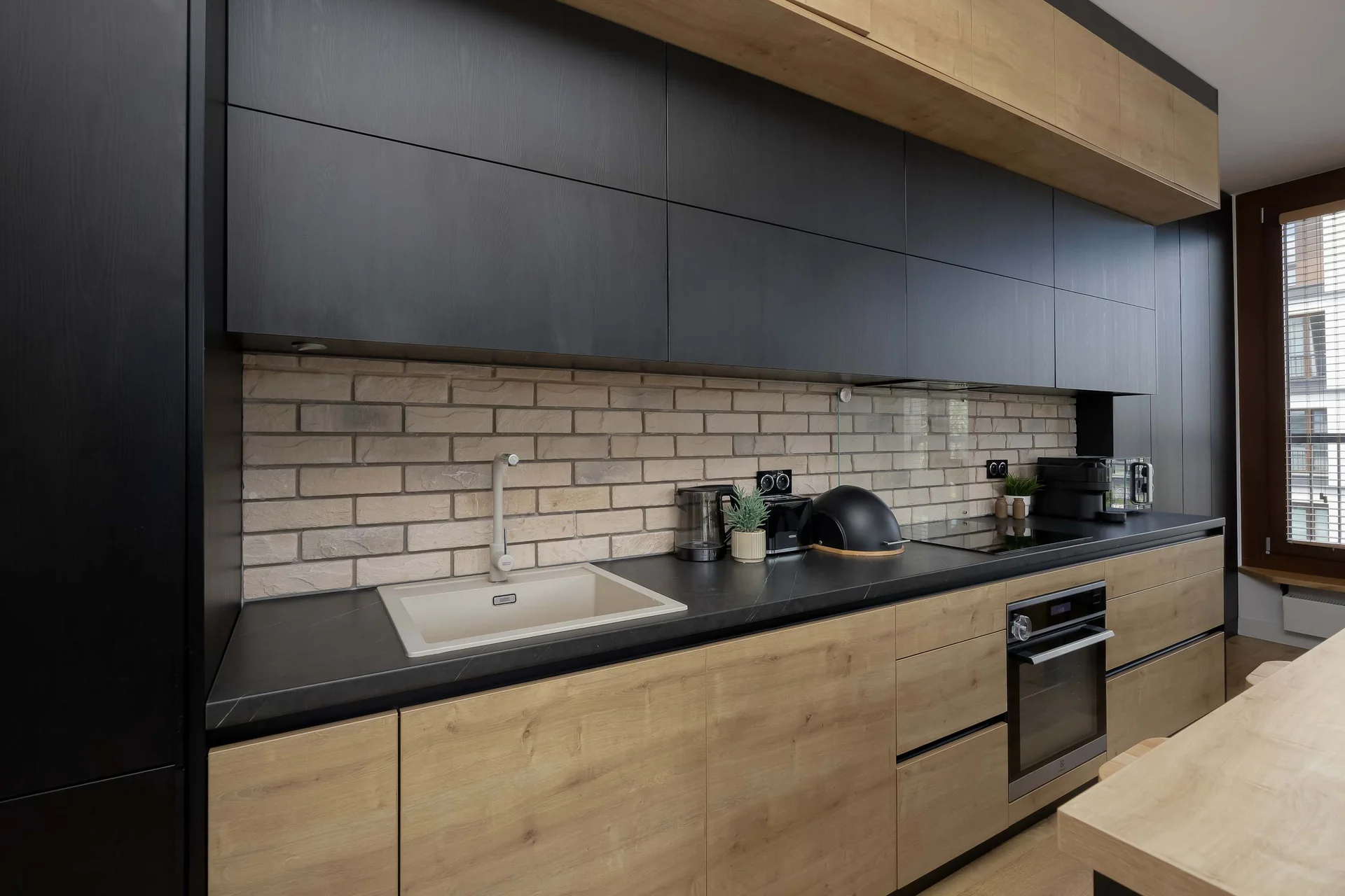 A close-up view highlights a modern kitchen workspace with matte black upper cabinets and warm wood lower cabinets. The white brick-patterned tile backsplash complements the dark countertop and built-in appliances.