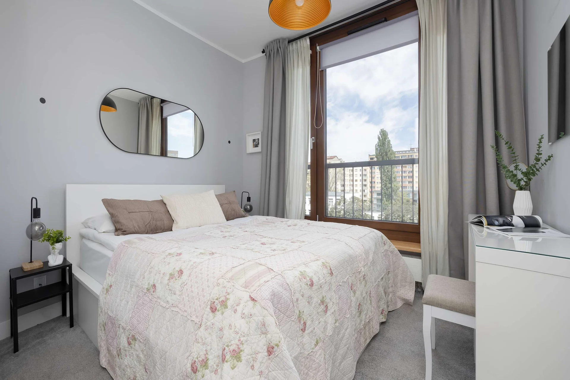 A bright bedroom is furnished with a double bed adorned in a light, patterned bedspread. A large window brings in natural light, flanked by grey curtains and a small white vanity table on the side.