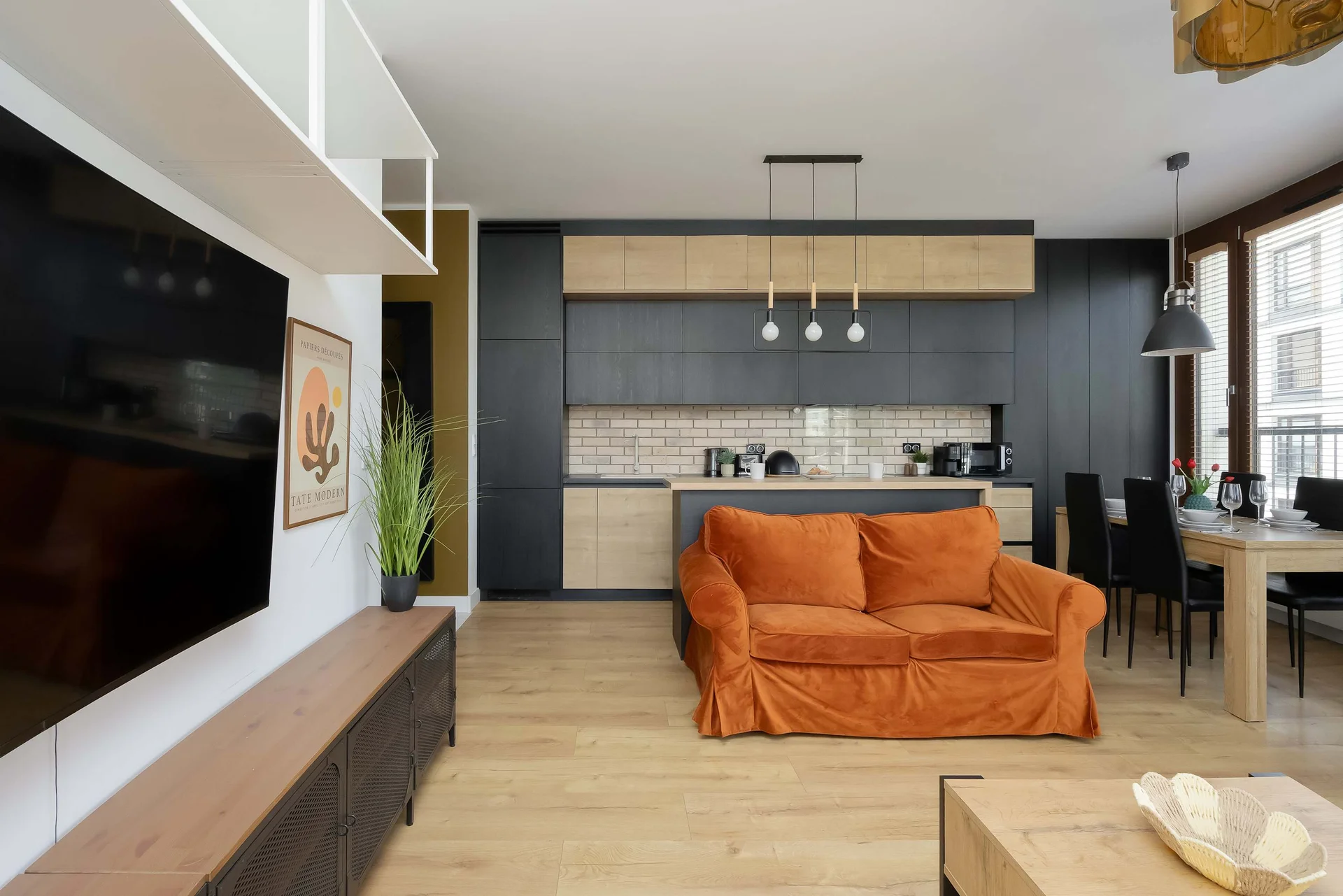 The living room perspective focuses on a wall-mounted television positioned above a long, low-profile media console. A partial view of the orange sofa and the adjoining kitchen space can be seen in the background.