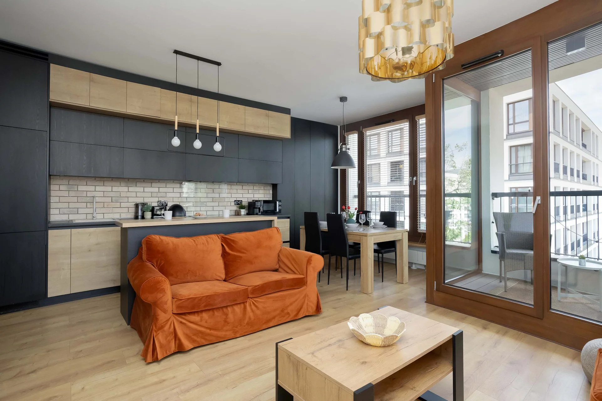 An open-concept living area features a vibrant orange sofa facing a wooden coffee table. The background opens into a sleek kitchen with dark cabinetry and a dining table, while large glass doors lead out to a balcony.