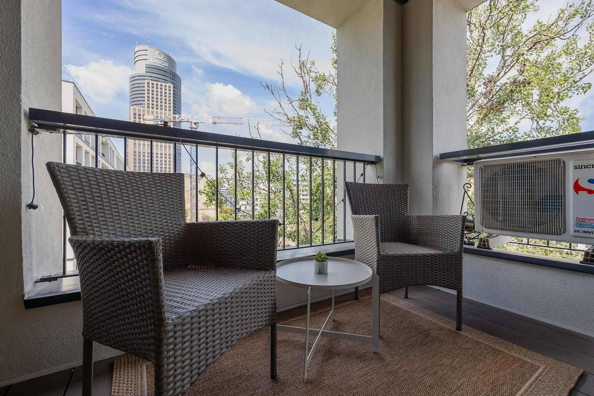 A cozy balcony space is arranged with two dark wicker armchairs and a small matching round table. The area is laid with a woven rug, offering a pleasant view of the surrounding trees and urban skyline.