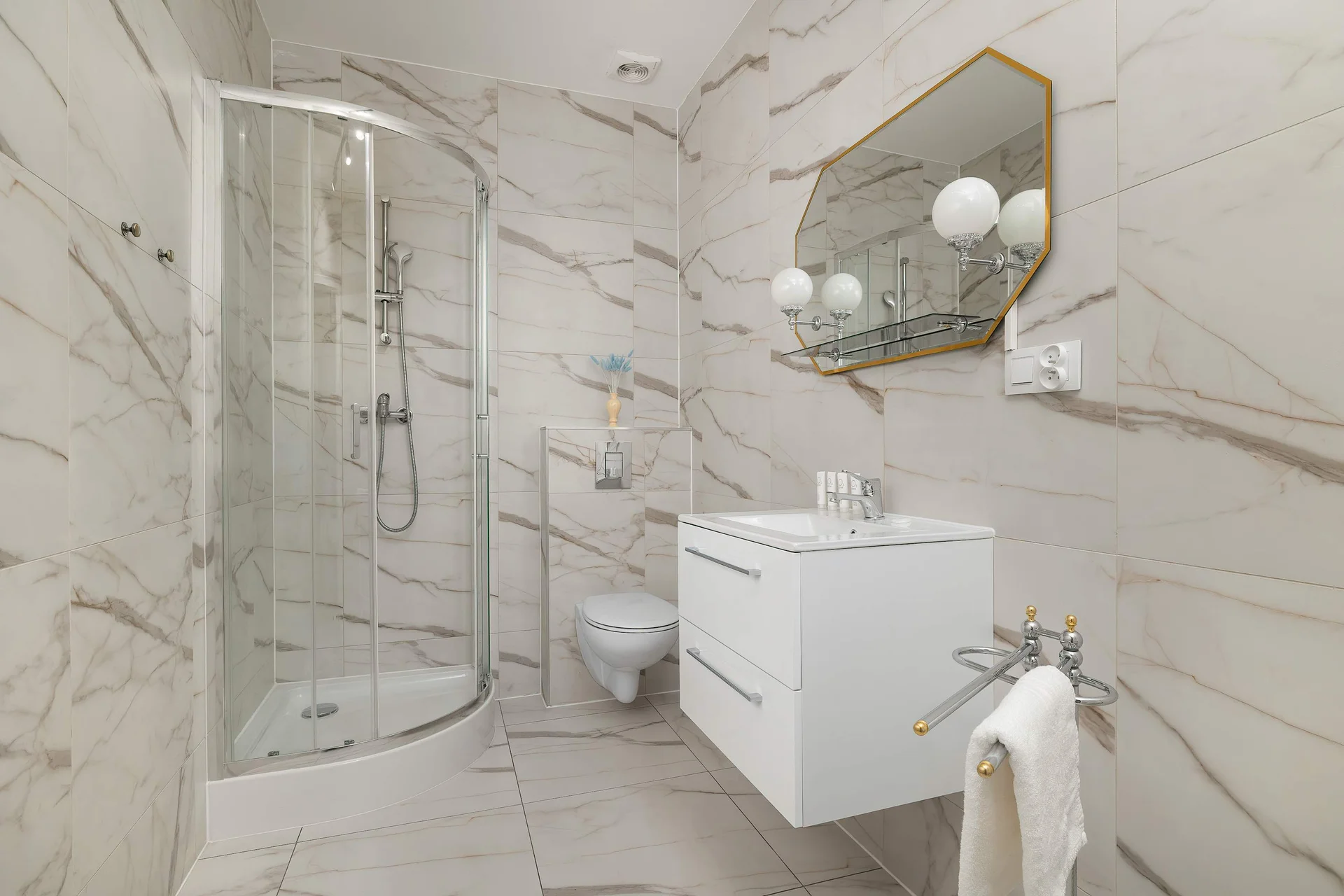 The pristine bathroom is fully lined with elegant white marble-patterned tiles and features a curved corner glass shower. A modern floating vanity with a rectangular mirror and soft decorative lighting completes the space.