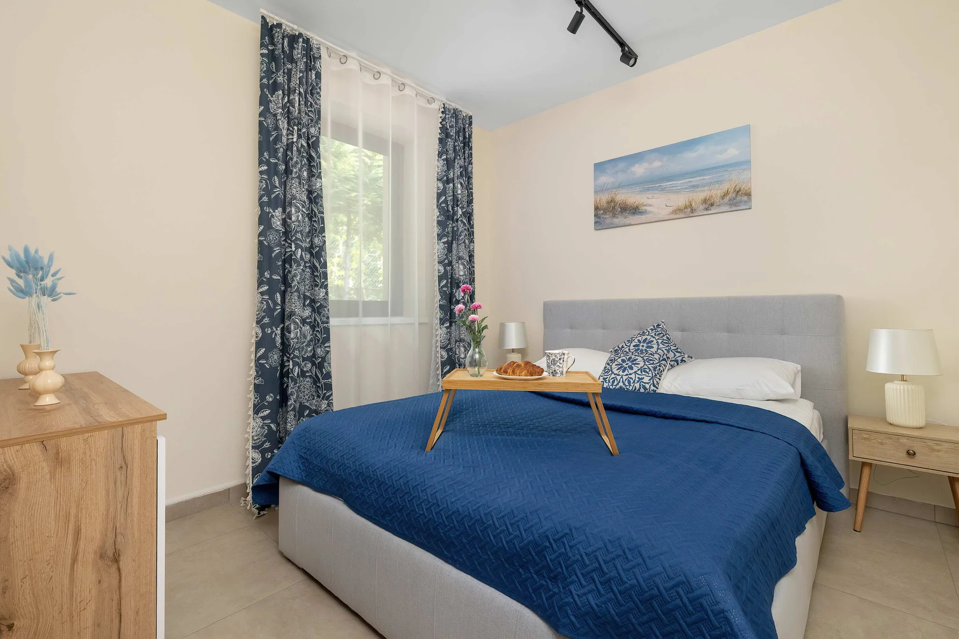 The tranquil bedroom is arranged with a comfortable double bed dressed in a deep blue quilted bedspread. A wooden breakfast tray rests on the mattress, and a large window with patterned blue and white curtains lets in natural light.