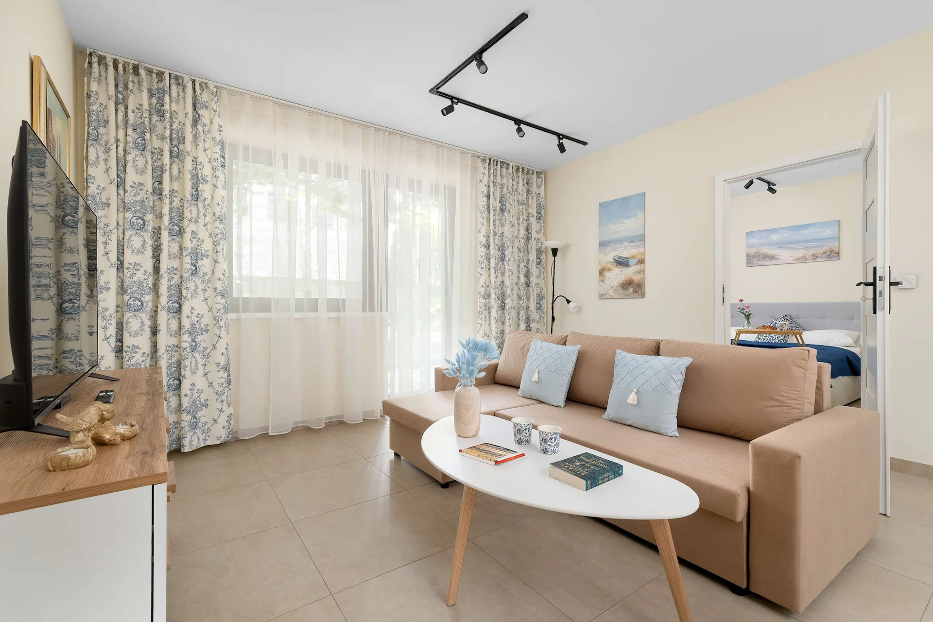 A cozy and bright living room features a comfortable beige sofa decorated with soft blue pillows and a sleek oval coffee table. Elegant floral curtains frame the window, while a flat-screen television sits on a white media console.