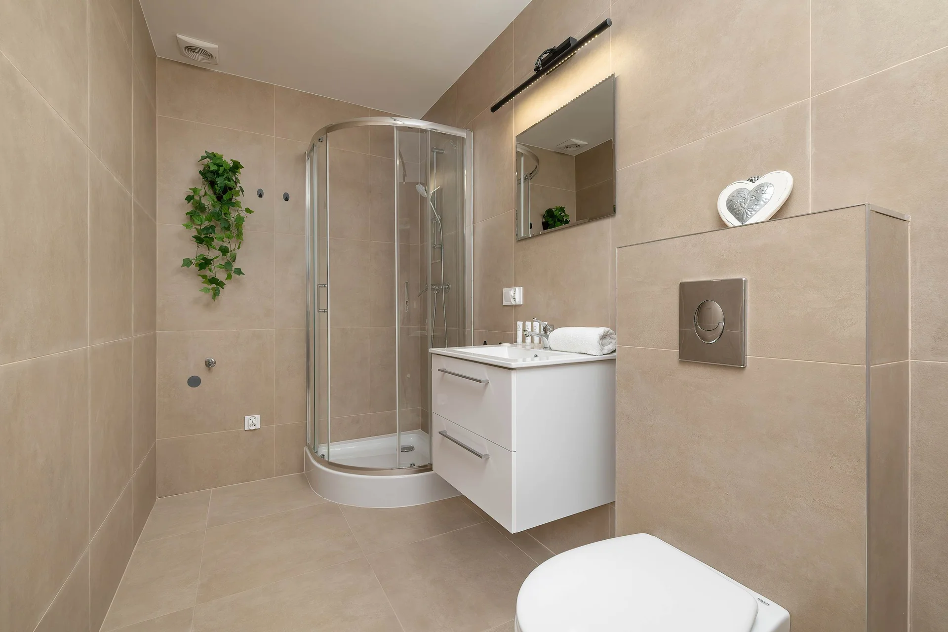 A bright bathroom features a glass corner shower cabin, a white vanity unit with a mirror, and neutral beige wall tiles. A small green plant hangs on the wall to soften the space.