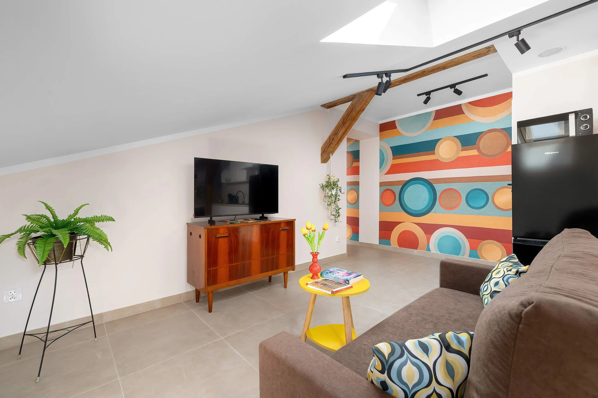 A spacious living room showcases a television on a vintage wooden cabinet, a brown sofa, and colorful geometric wallpaper in the background. Potted green plants add life to the room.