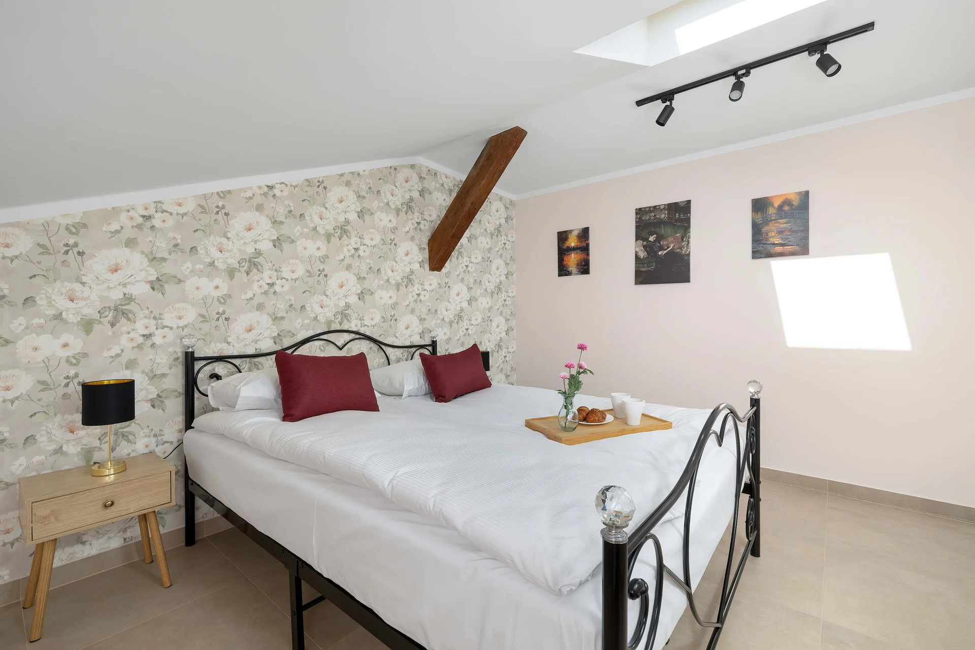 A stylish bedroom features a bed with white bedding and red pillows, positioned against a floral patterned wallpaper accent wall. Small wooden nightstands flank the bed on either side.