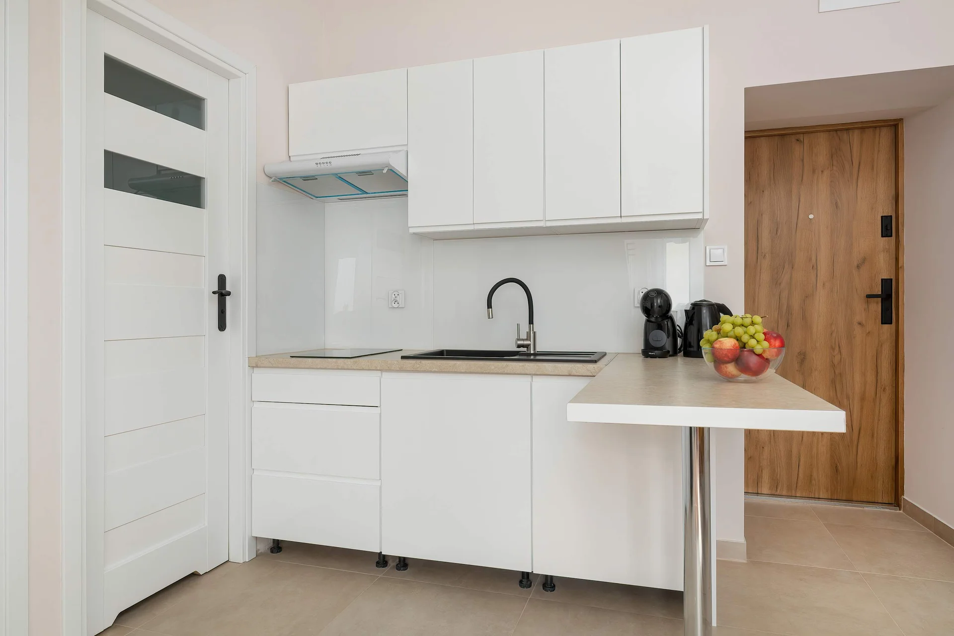A clean, white kitchenette is visible next to an open doorway, showcasing a minimalist design and a black countertop appliance. The flooring consists of light-colored, glossy tiles.