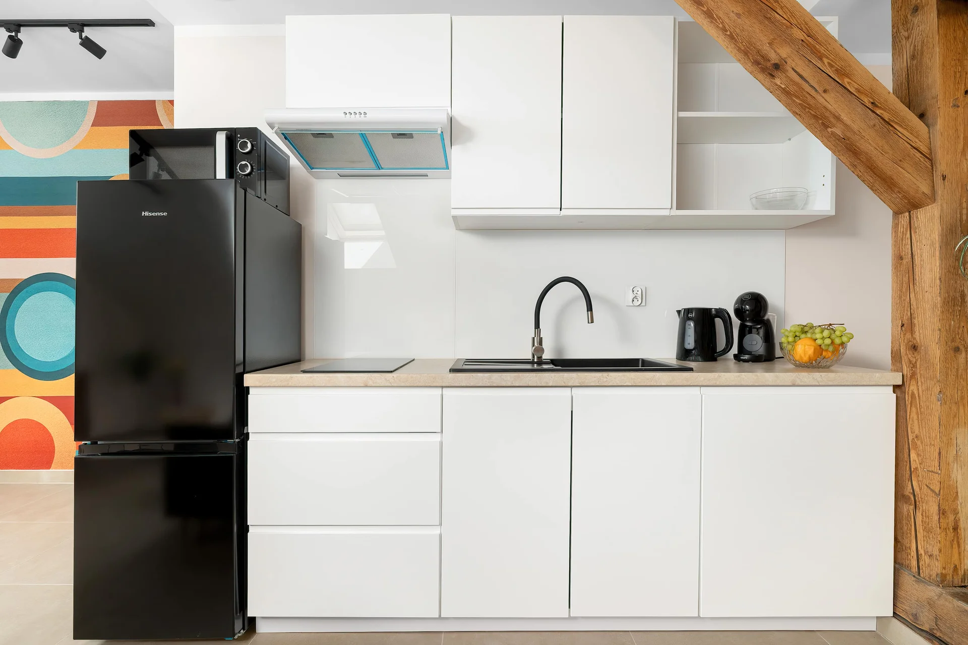 A compact white kitchenette is equipped with a black refrigerator, a sink, and built-in cabinetry. An exposed wooden beam on the right adds a rustic touch to the modern setup.