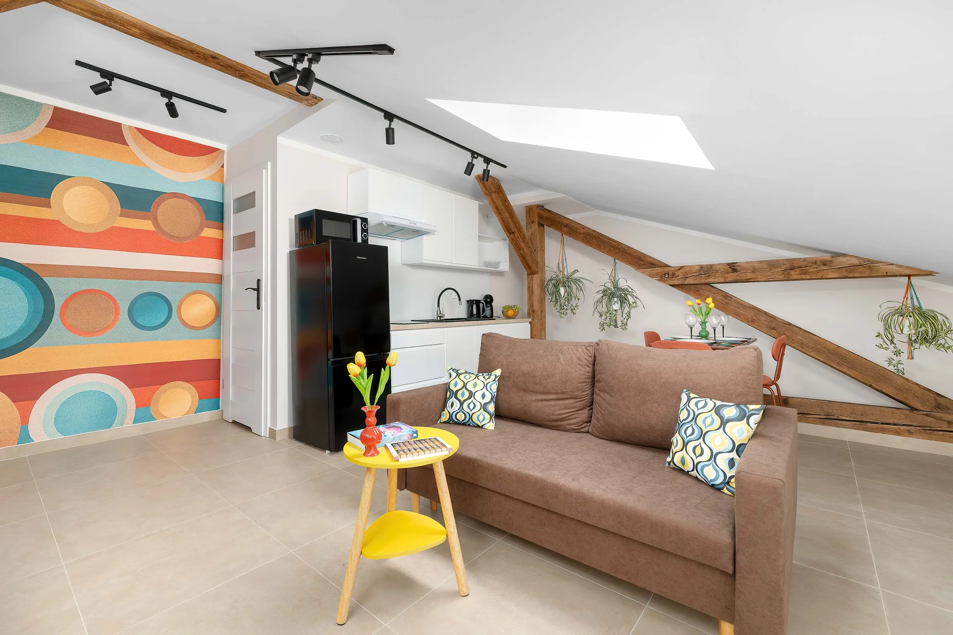A view of the living area highlights a brown sofa, a small yellow side table, and vibrant, multi-colored circular pattern wallpaper. Exposed wooden ceiling beams run across the top of the room.