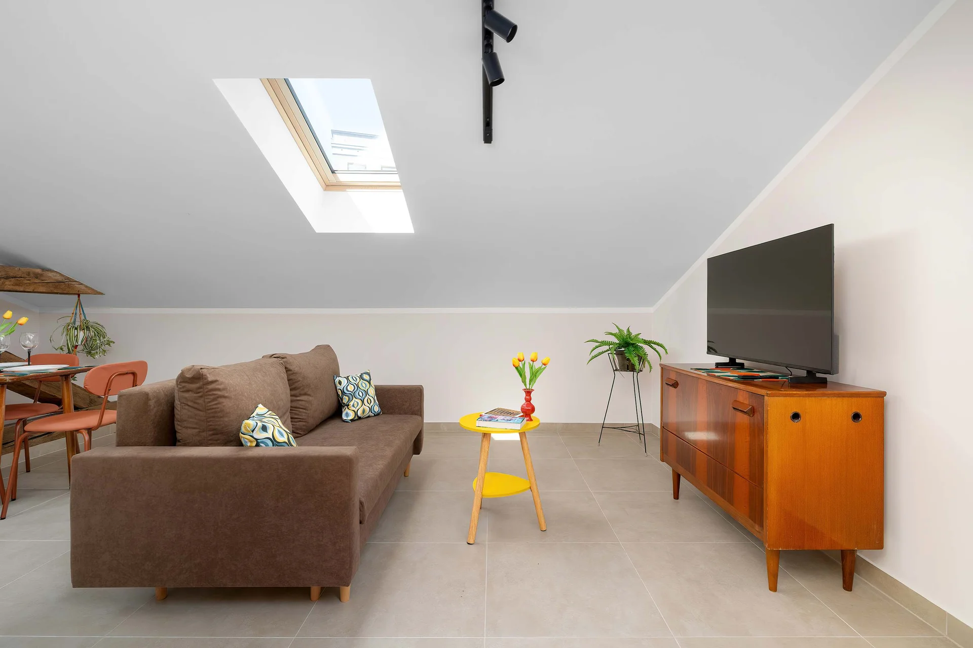 An open-plan living room contains a brown sofa, a wooden TV stand, and a skylight window illuminating the space. A small yellow coffee table sits in the center, adding a pop of color.