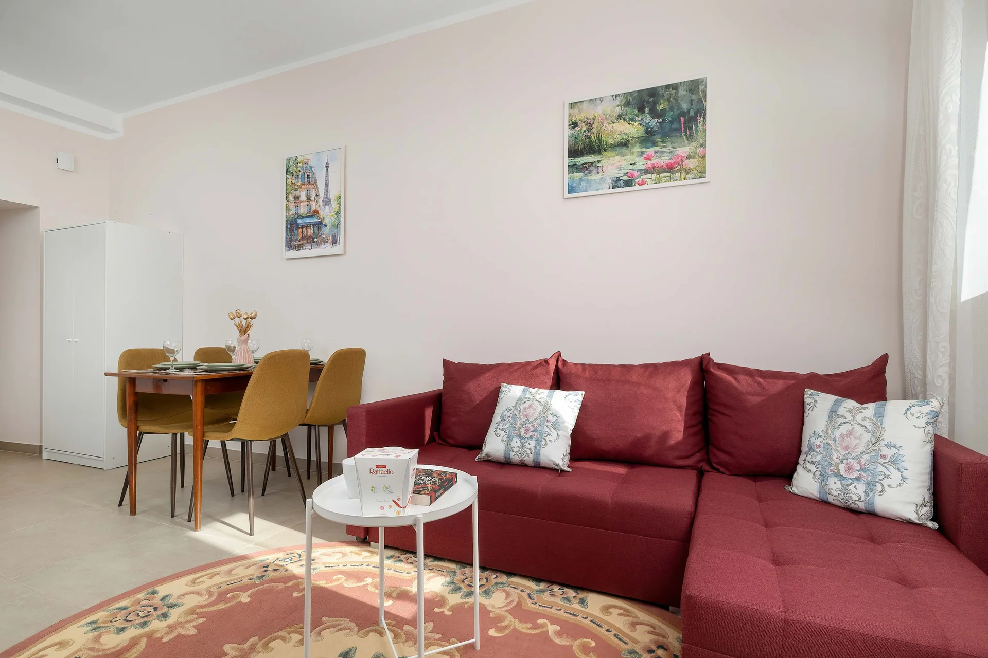 A dining area transitions into a living space, featuring a wooden table with chairs and a plush red sofa on a patterned rug. Two small paintings hang on the wall, enhancing the decor.