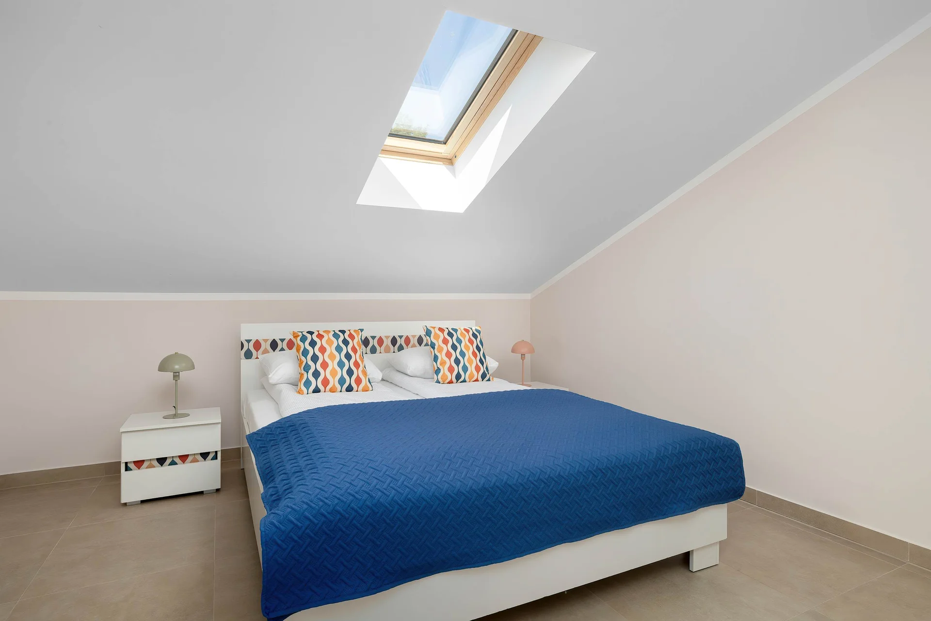 A bright bedroom offers a large double bed with a vibrant blue blanket under a slanted ceiling with a skylight window. A small white bedside table sits next to the bed, creating a clean and minimalist look.