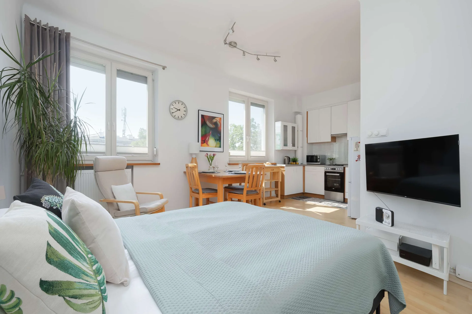 Another perspective showcases the bed, TV, and dining area within the open-plan studio. Light walls and natural wood flooring enhance the airy feel.