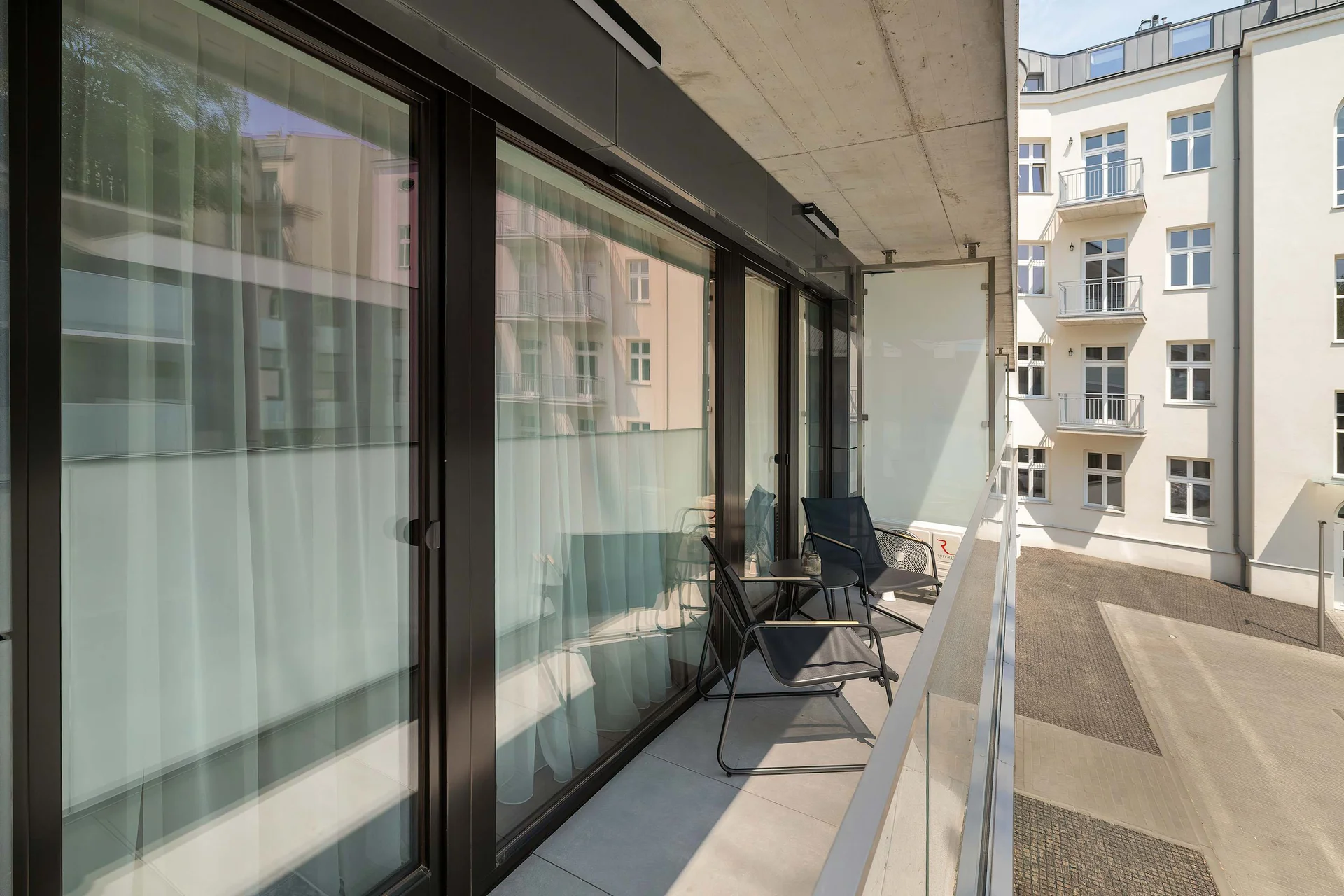 A long, private balcony is enclosed by modern glass and dark metal railings. The outdoor area overlooks a clean, paved inner courtyard between contemporary buildings.