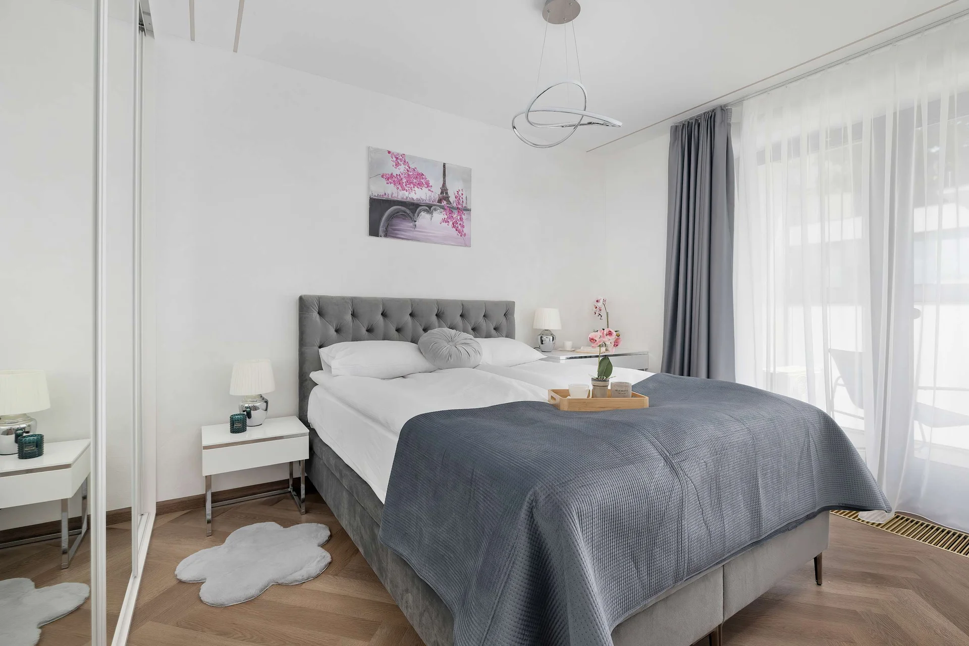A stylish bedroom offers a large double bed dressed in elegant grey and white linens. Sun streams through large windows dressed with delicate sheer curtains, illuminating the entire space.