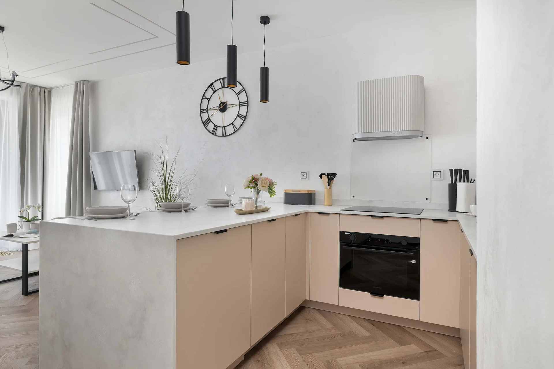 An elegant kitchenette boasts light wooden lower cabinetry paired with seamless white countertops. Modern integrated appliances and stylish black pendant lights complete the functional cooking space.
