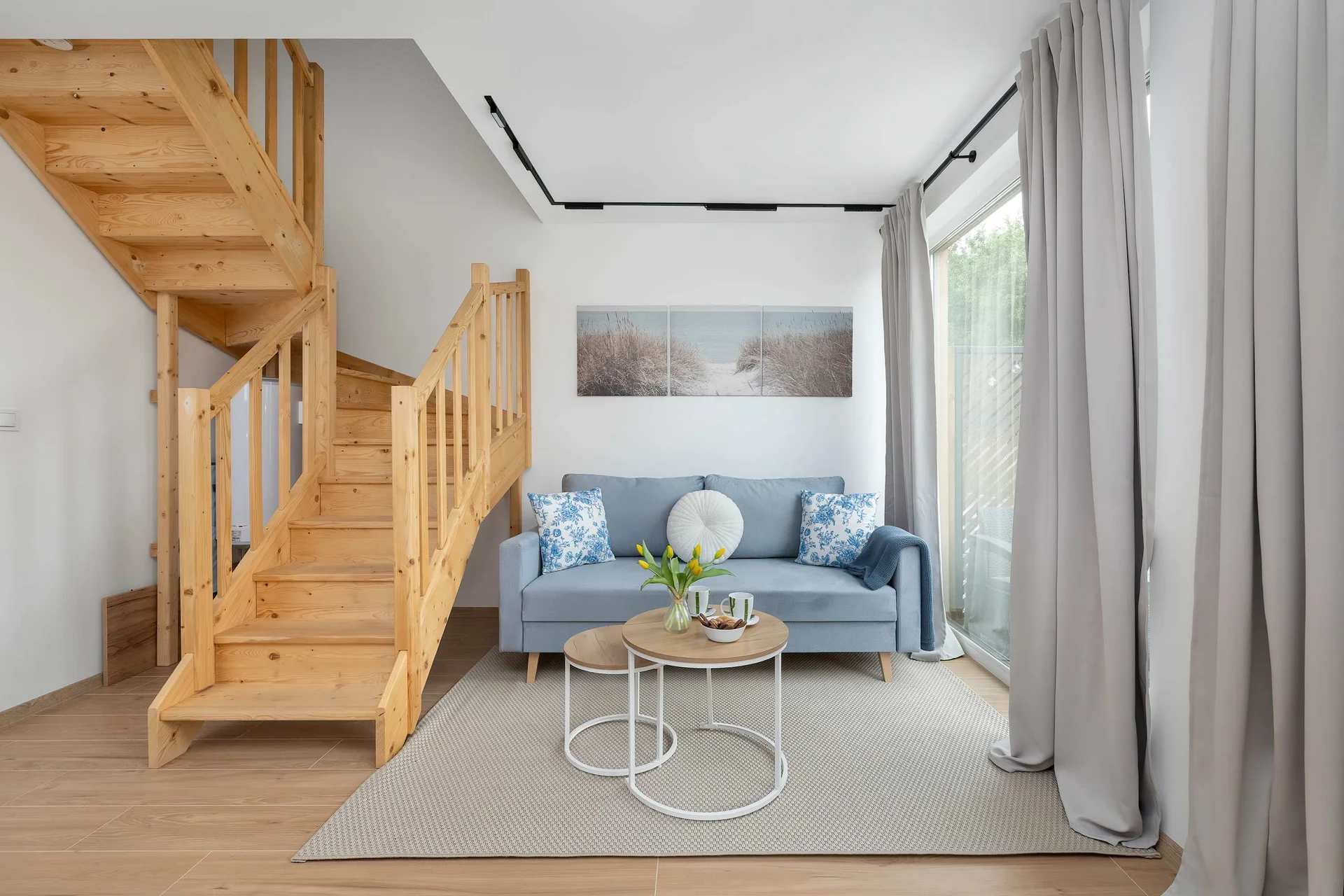 A modern living area showcasing a wooden staircase leading upstairs and a comfortable seating arrangement below. A small table rests on a neutral area rug in the center of the room.