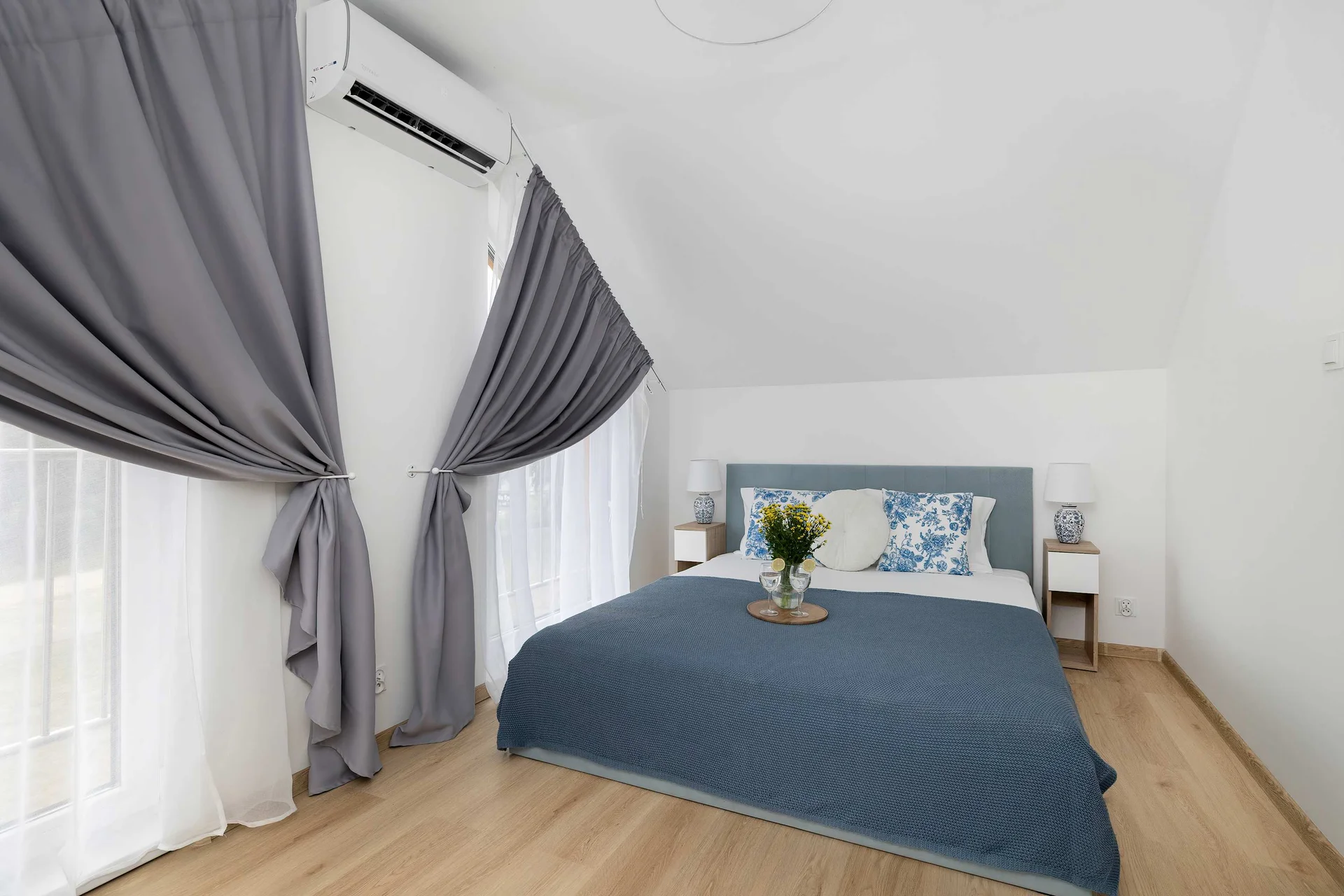A brightly lit bedroom with a large double bed dressed in blue linens under a slanted ceiling. Soft curtains frame the window, enhancing the peaceful atmosphere.