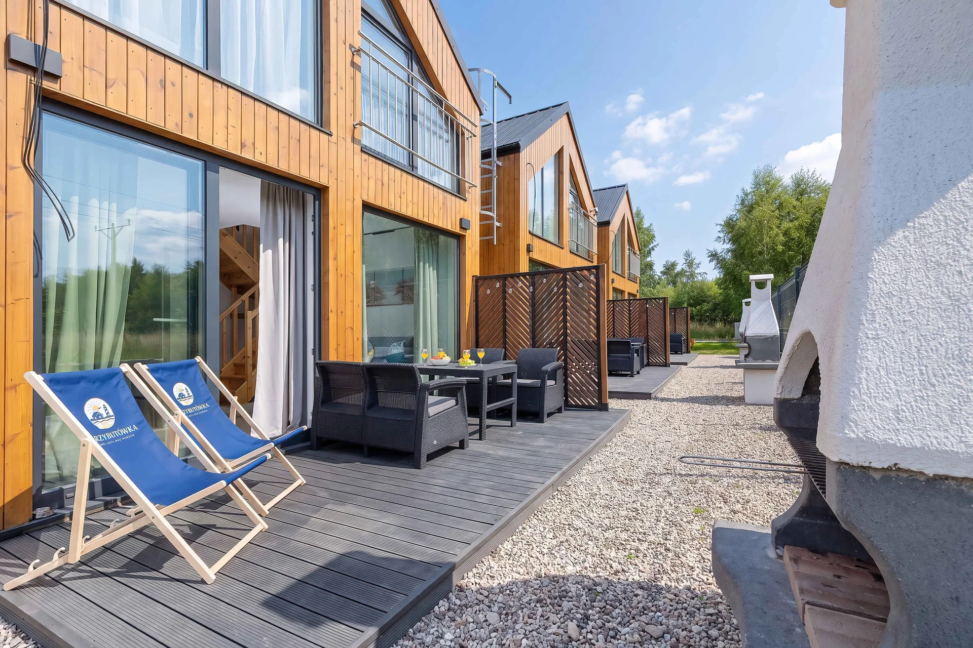 An outdoor terrace view of modern wooden cottages with blue deck chairs set out on the paved ground. The open doors hint at a seamless transition between indoor and outdoor living.