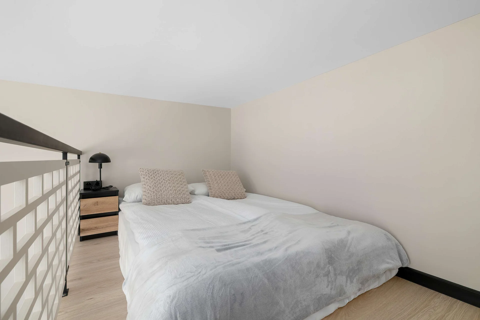 A cozy loft bedroom is furnished with a double bed covered in white linens and neutral-toned pillows. A low brick wall serves as a safety barrier overlooking the lower level of the apartment.