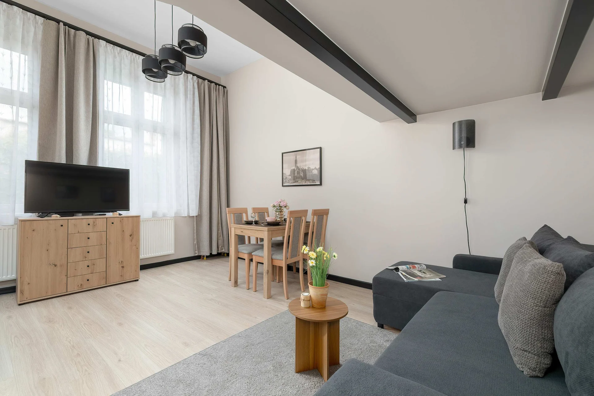 A spacious living room features a dark grey corner sofa paired with a small wooden coffee table. A wooden dining table sits in the background near a large window, while a TV unit is positioned opposite the seating area.