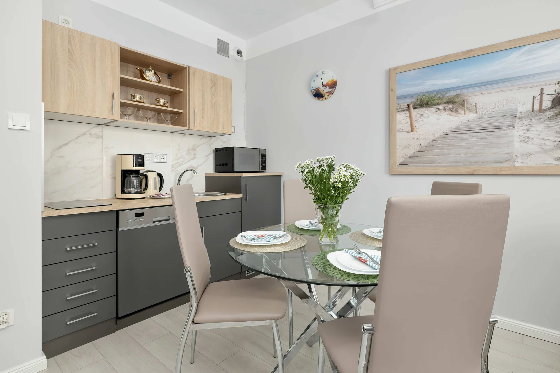A kitchen is equipped with grey cabinets, light wood uppers, and a glass dining table. A beach landscape painting hangs on the wall.