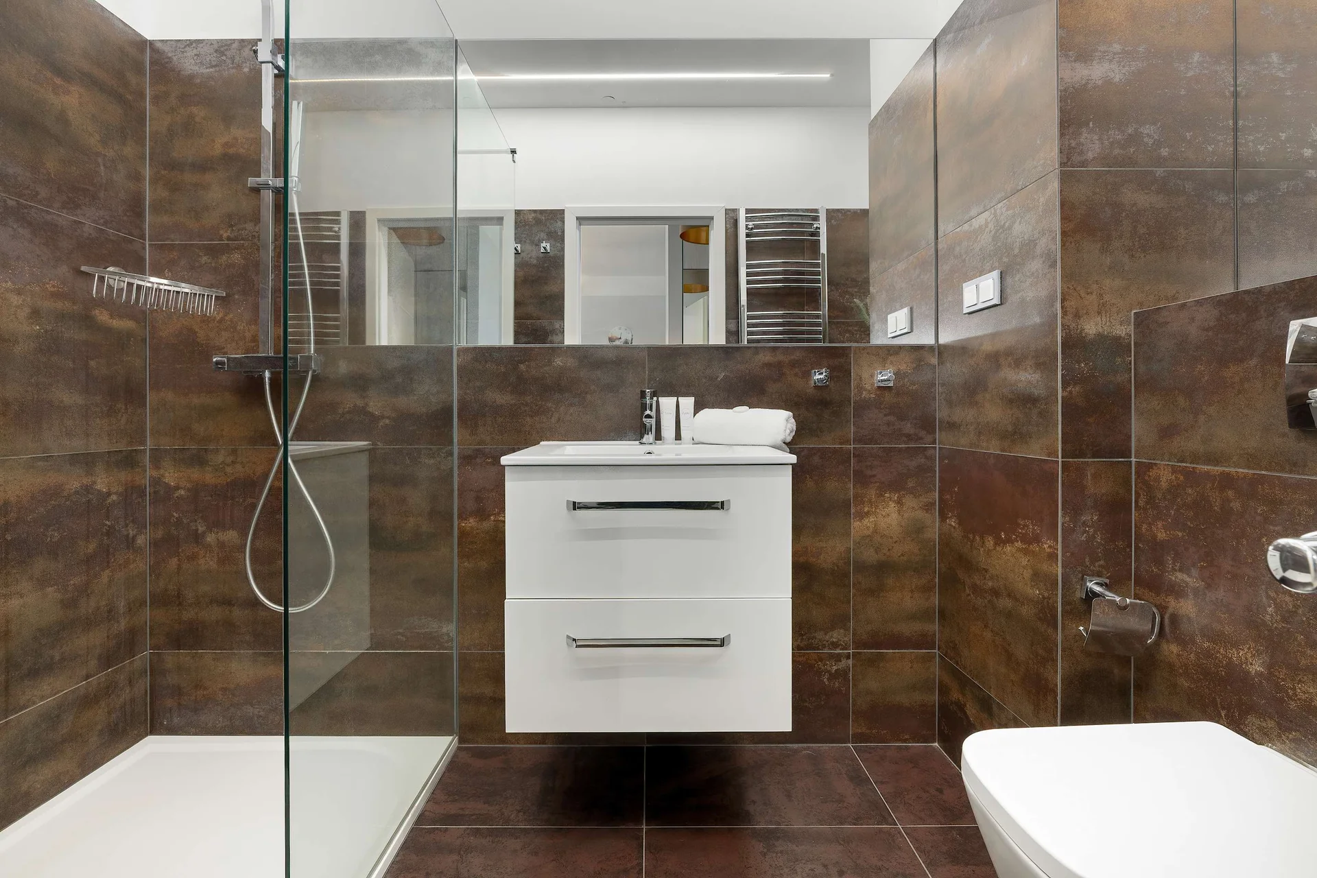 A bathroom features brown marble-textured tiles and a white vanity unit with drawers. A glass partition encloses the walk-in shower.