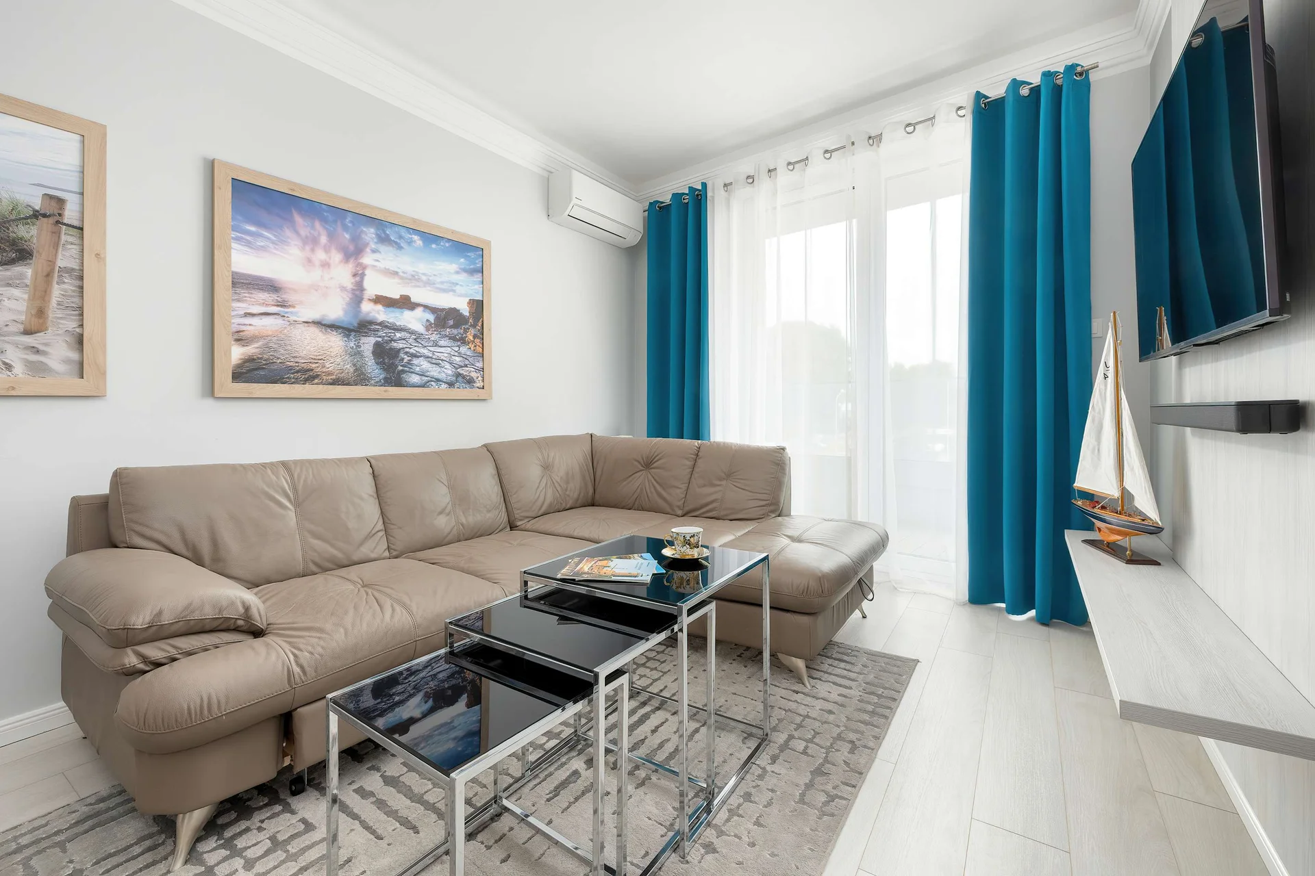 A living room features a beige leather corner sofa, glass coffee tables, and a wall-mounted TV. Vibrant blue curtains flank the window.