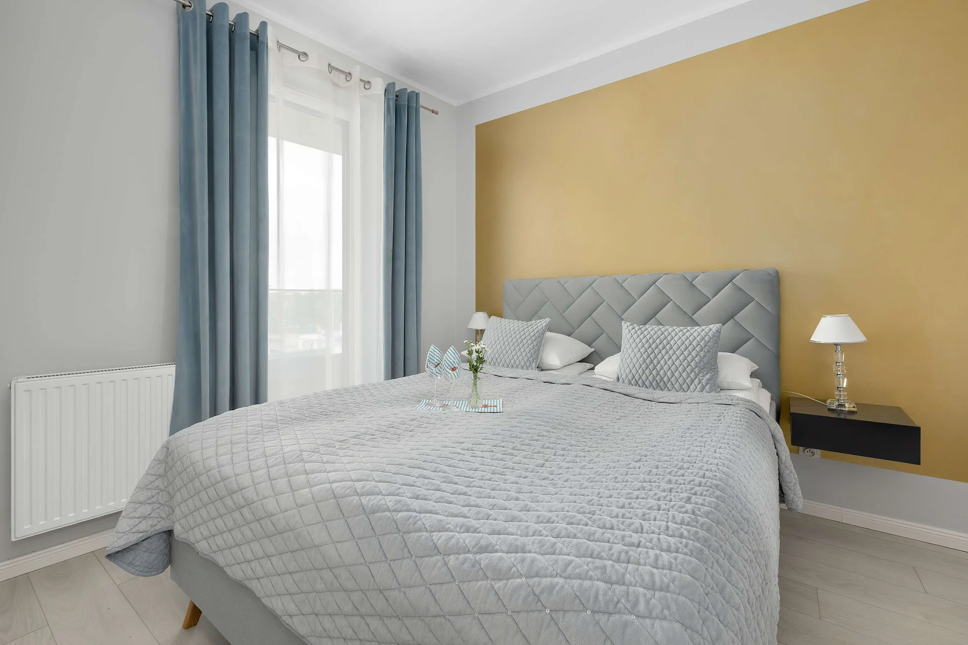 The bedroom showcases a double bed with a grey tufted headboard against a warm yellow accent wall. Blue curtains frame the large window.