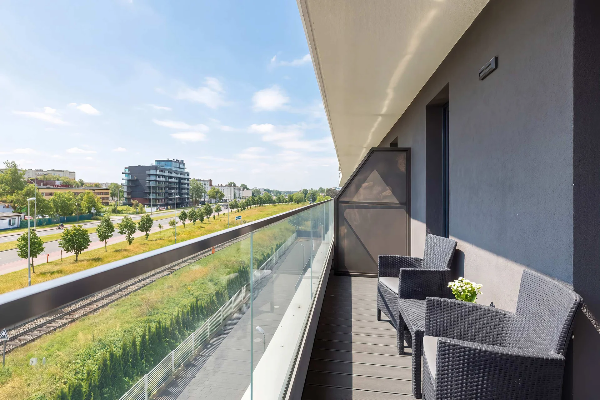 A long balcony with a glass railing is furnished with dark woven chairs. The view looks out onto a green corridor and train tracks.
