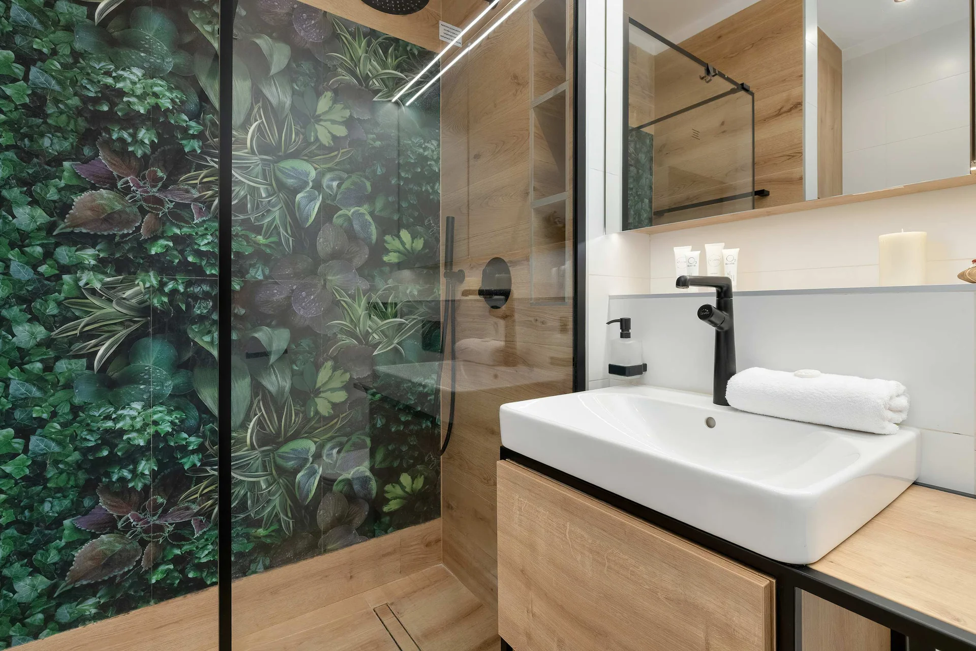A modern bathroom vanity setup displays a rectangular white sink resting on a light wood counter with a black faucet. A vibrant green foliage wall panel is visible behind the glass shower screen, adding a natural touch.