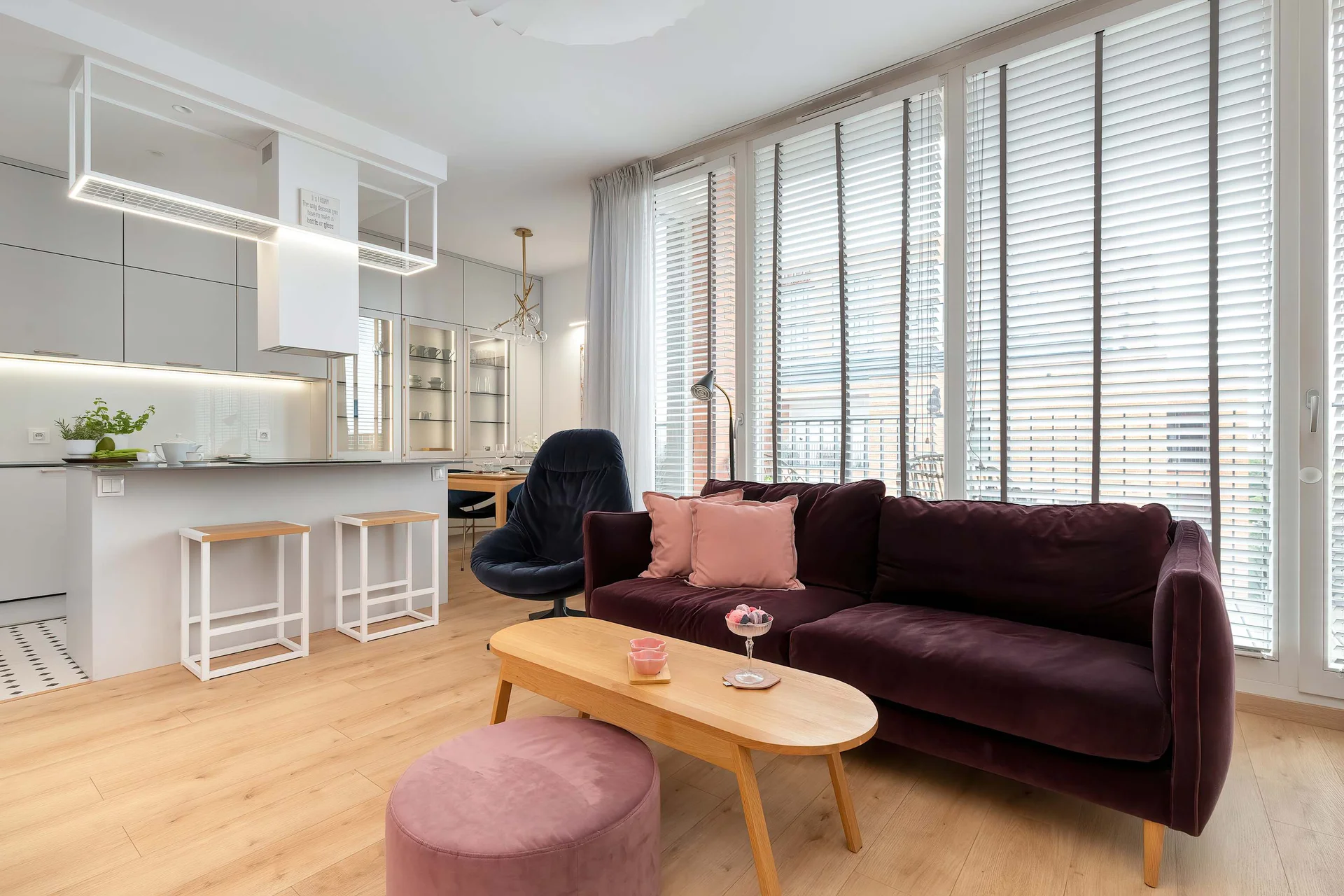 An open-plan living room and kitchen area showcases a dark purple sofa, a wooden coffee table, and large windows with horizontal blinds. Modern white kitchen cabinets and a breakfast bar with stools are visible in the background, creating an airy fee