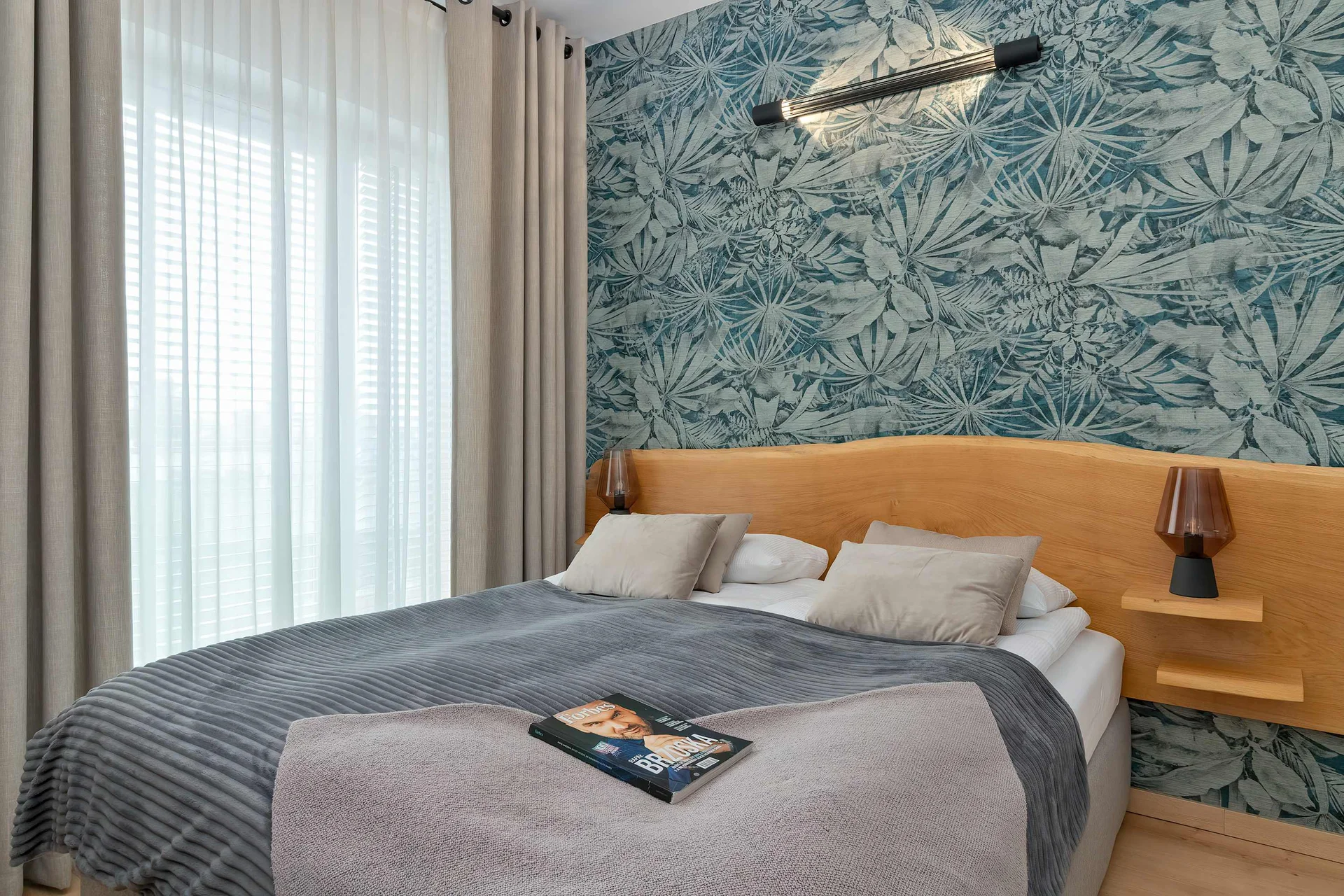 A comfortable double bed is dressed in grey and white linens beneath a distinctive blue botanical wallpaper pattern. A wooden headboard and small matching bedside tables frame the sleeping area symmetrically.