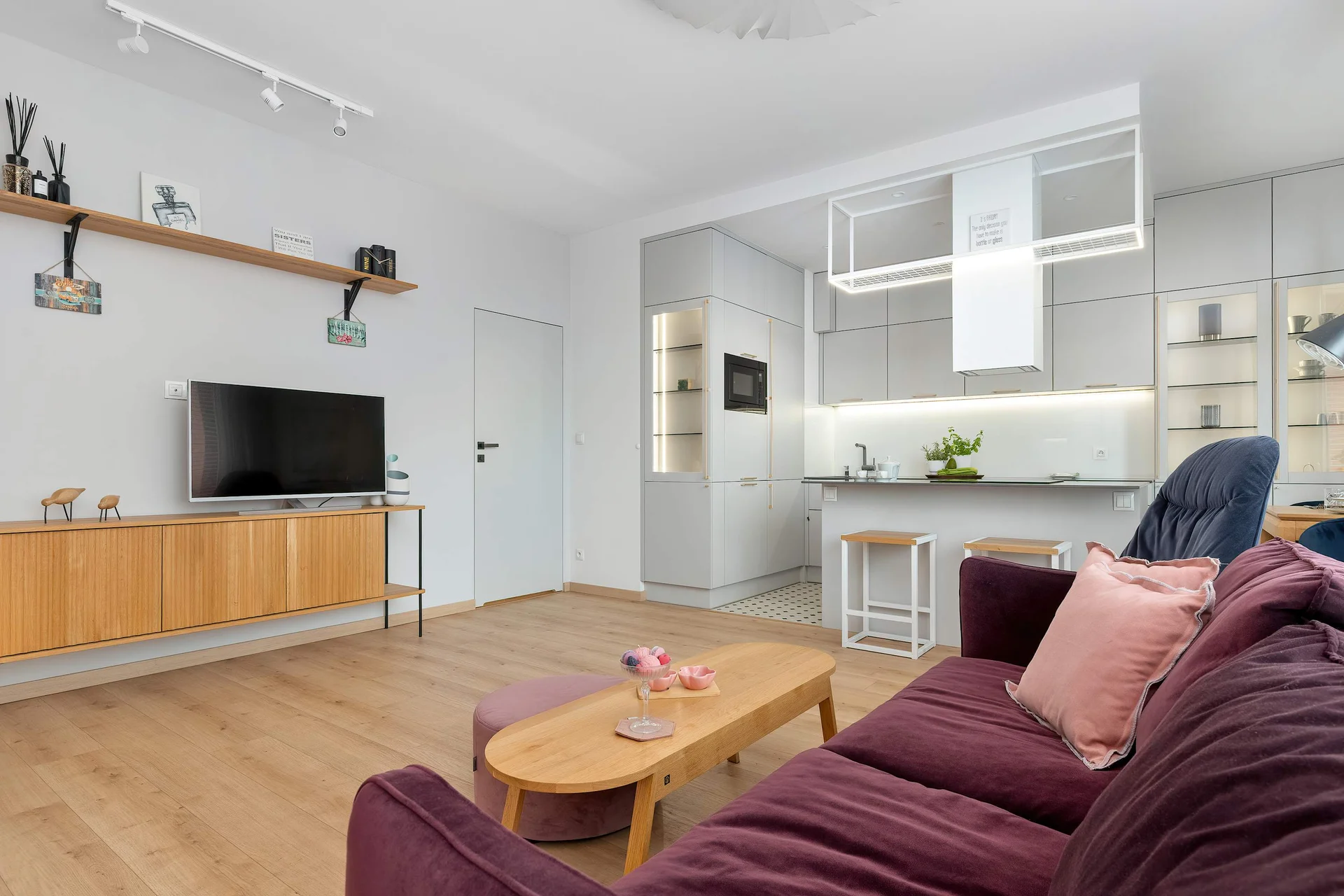 A spacious living room layout centers around a wall-mounted flat-screen TV over a long, low-profile wooden media console. A plush dark purple sofa and a light wooden coffee table occupy the foreground under bright overhead lighting.