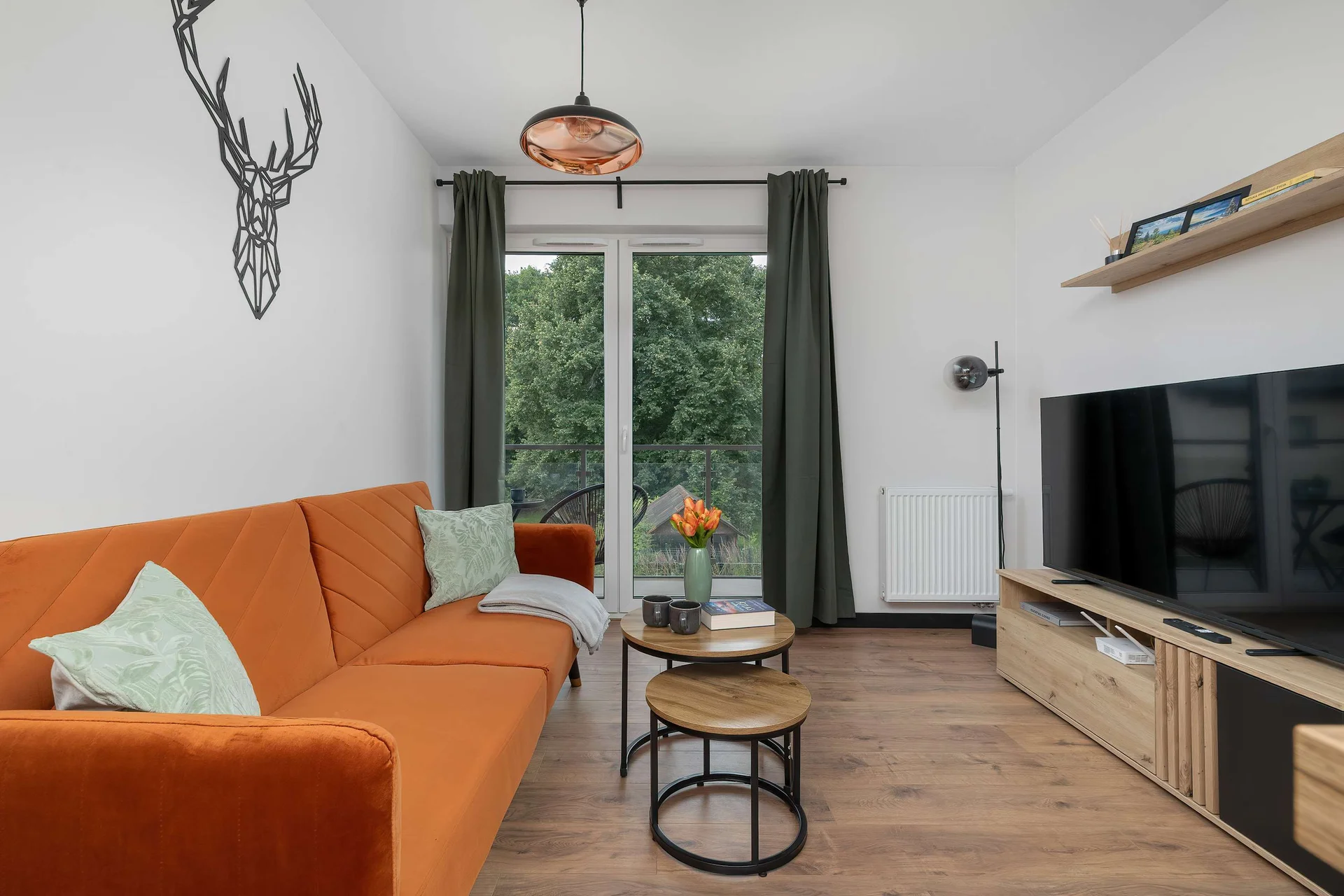 A modern living room features a sofa placed opposite a wooden media console. Large glass doors leading to a balcony allow natural light to fill the space, creating a bright atmosphere.