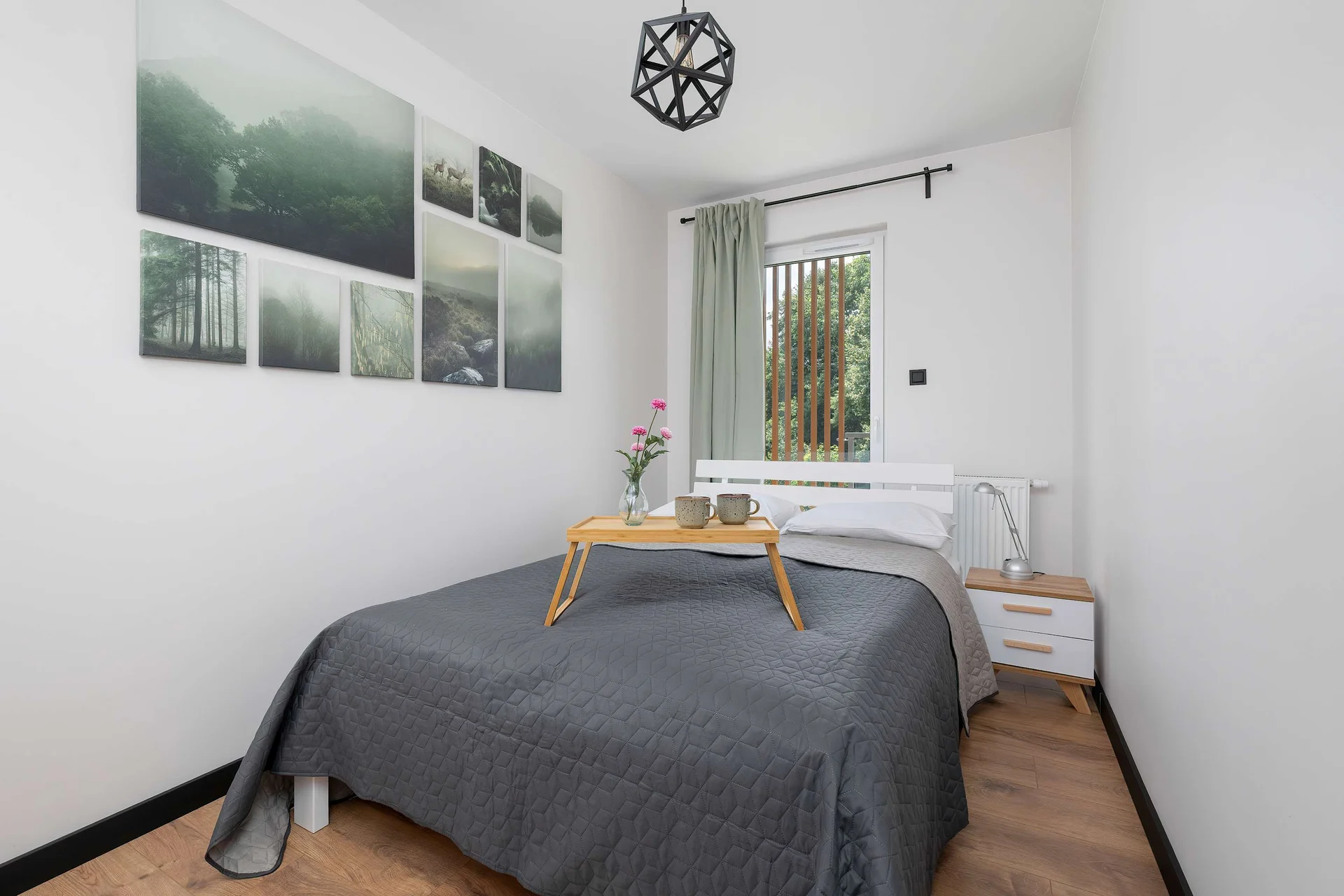A neat bedroom contains a double bed with a grey bedspread. Art pieces hang symmetrically on the crisp white wall behind the bed.